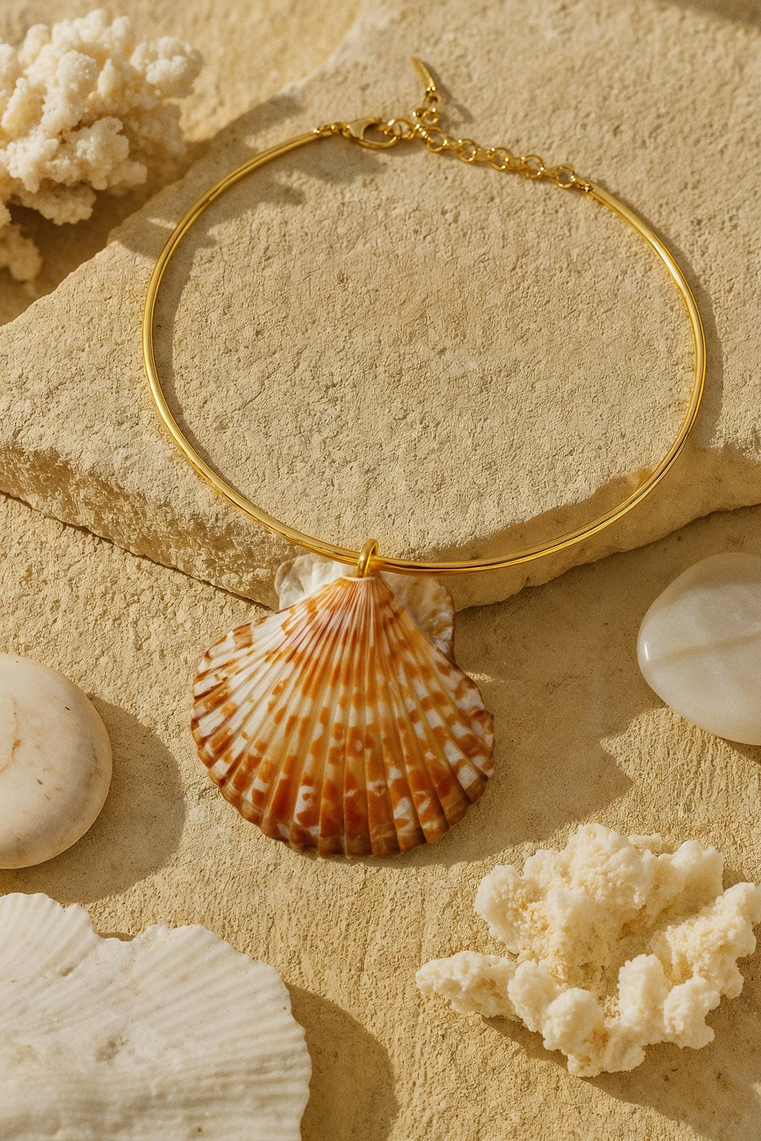 Gold necklace with a shell pendant displayed on a sandy surface, surrounded by seashells, coral, and white stones in a beach-themed arrangement.