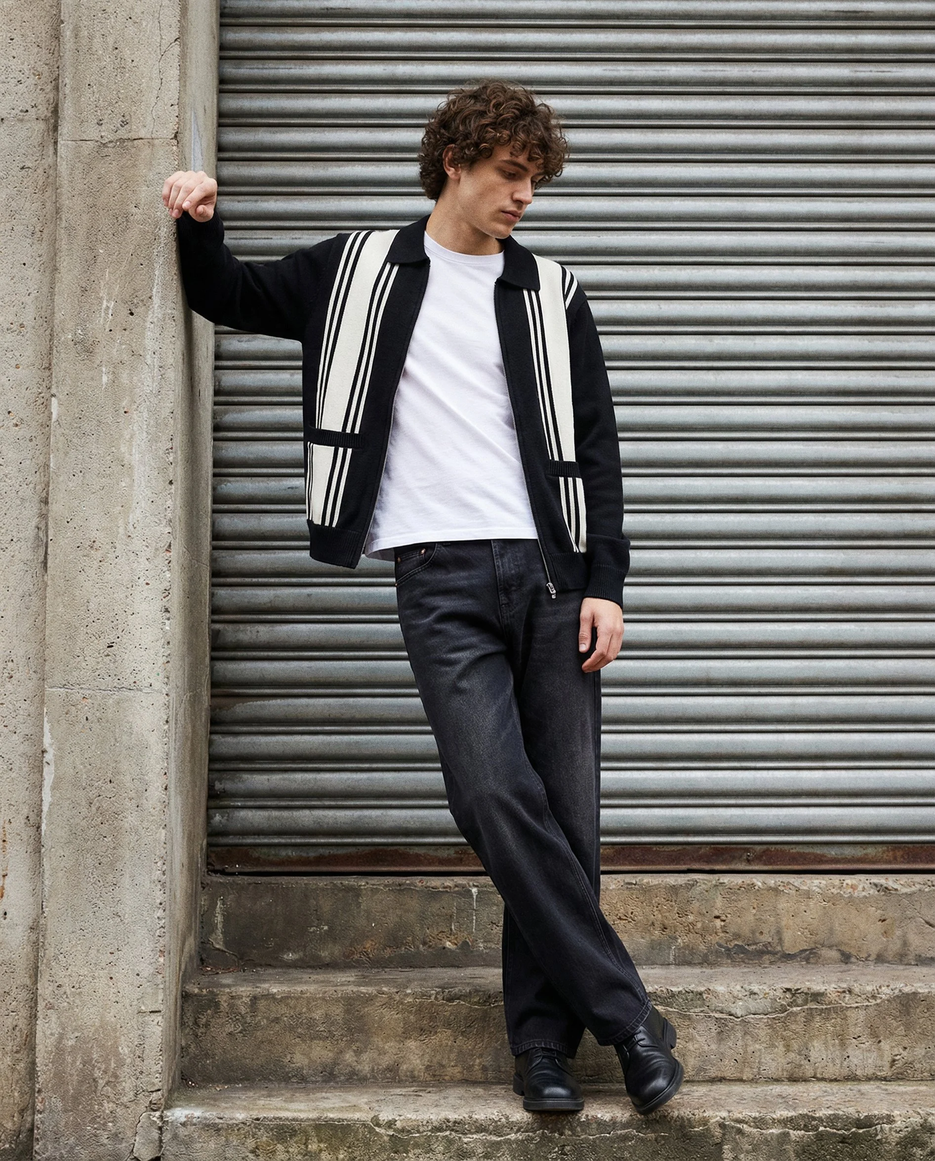 Young man with curly hair wearing a black and white jacket, white t-shirt, dark jeans, and black boots, standing against a concrete wall and metal roll-up door.