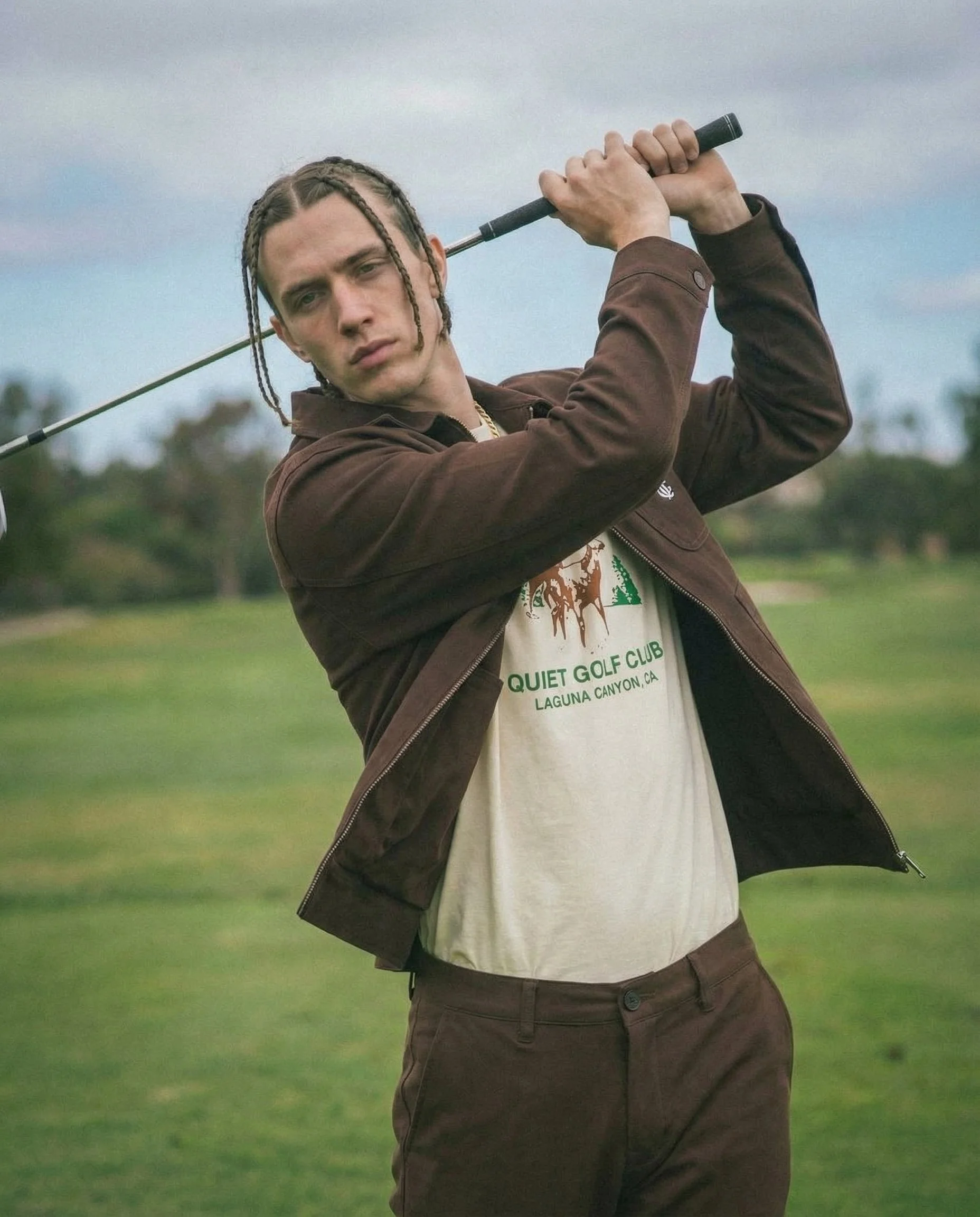 A young man with braided hair playing golf outdoors on a cloudy day, wearing a brown jacket over a white shirt with a golf course background.