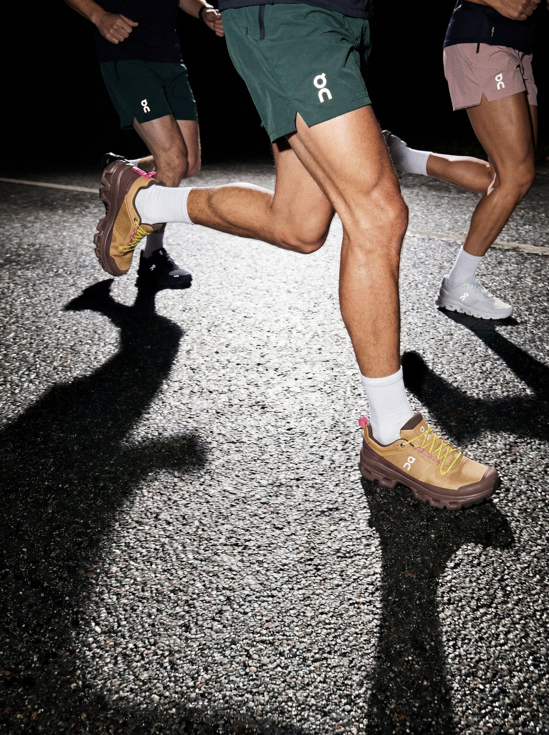 Close-up of three runners' legs and feet running on a gravel road at night, wearing athletic shoes and shorts, with shadows cast on the ground by bright lighting.