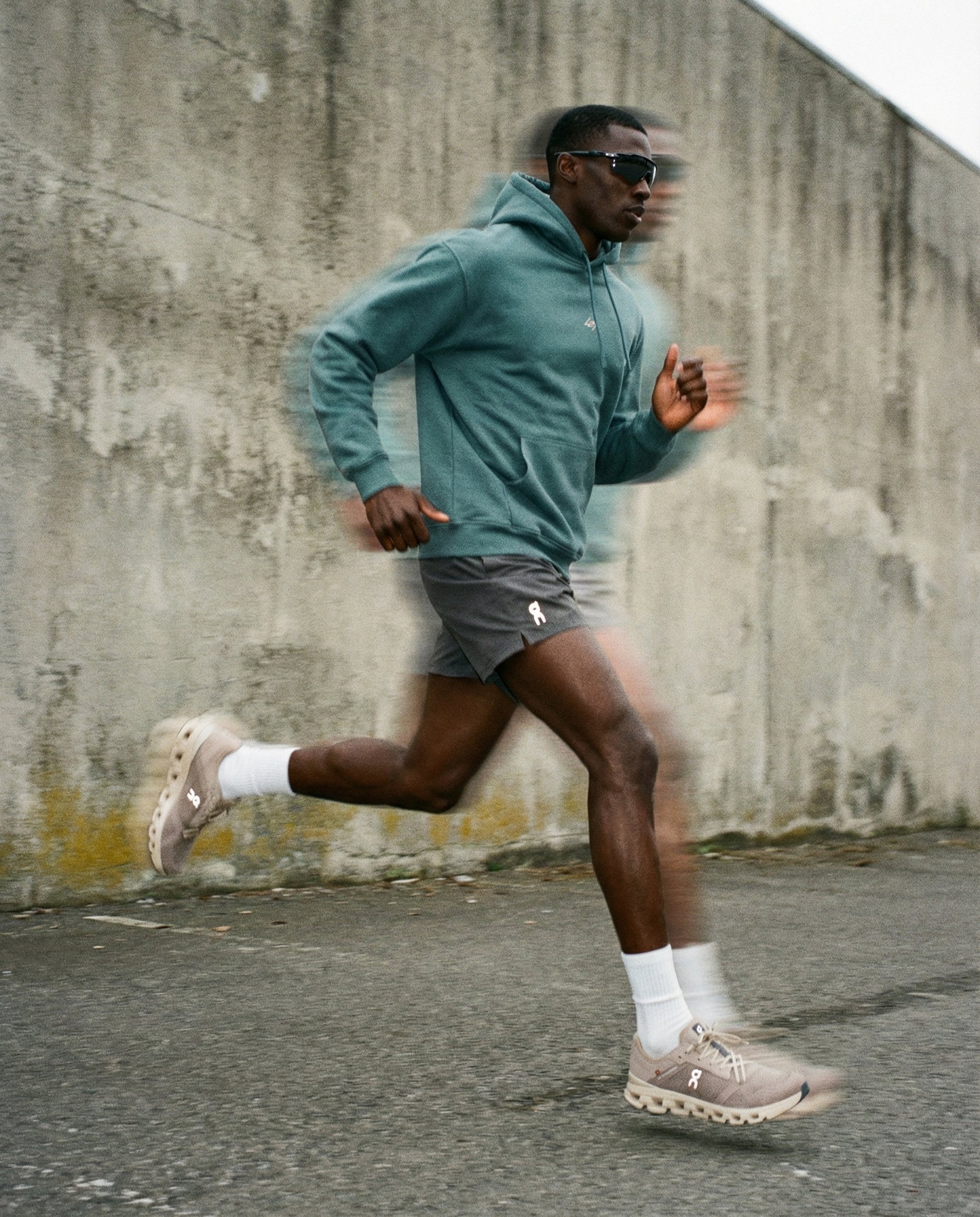 A man jogging outdoors on a concrete pavement against a weathered concrete wall, wearing a teal hoodie, dark shorts, beige sneakers, white socks, and dark sunglasses.