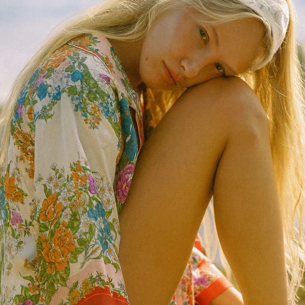 A young woman with long blonde hair resting her head on her knee, wearing a headband and a colorful floral shirt, outdoors in natural light.
