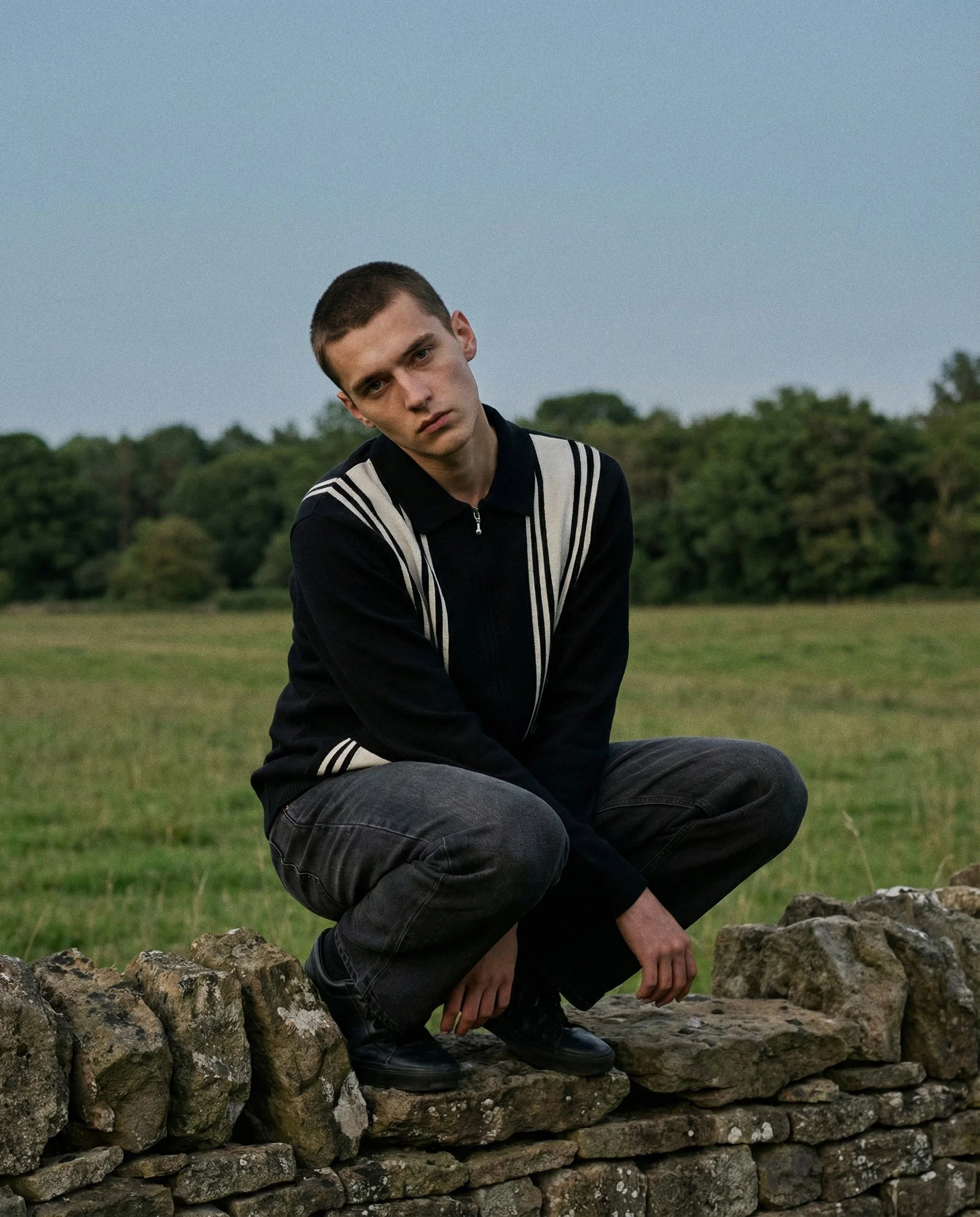 A young man with short hair and a serious expression, squatting on a stone wall outdoors, wearing a black and white jacket and black shoes, with a green landscape and trees in the background.