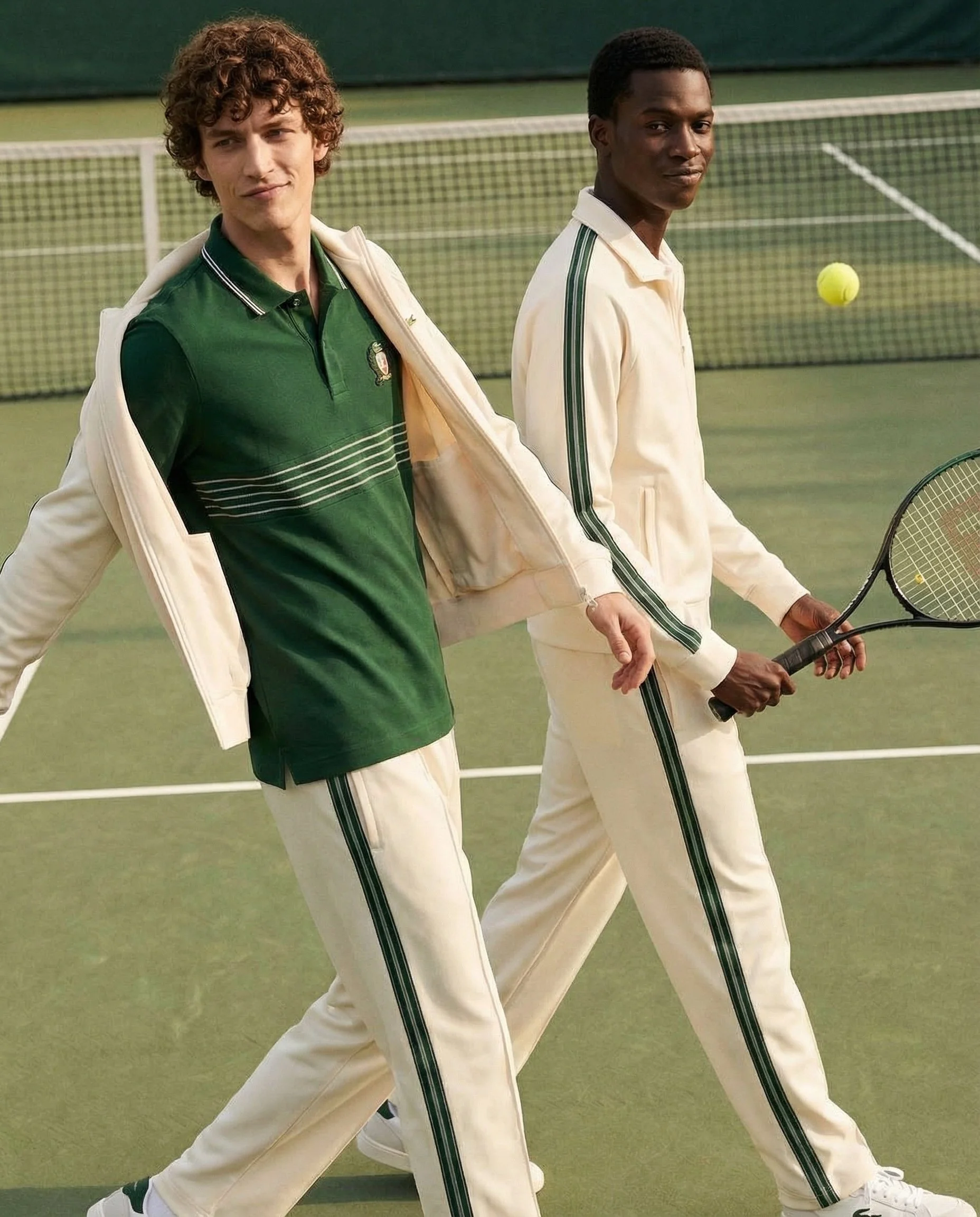 Two young men walking on a tennis court with tennis racquets, dressed in vintage sportswear, with a tennis ball in the air.