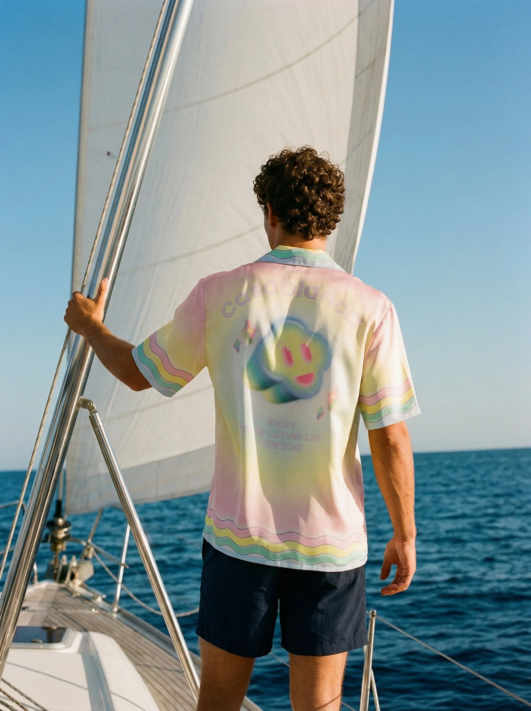 A person with curly hair wearing a colorful t-shirt and navy shorts standing on a sailboat, holding onto a metal rail, with the ocean and blue sky in the background.