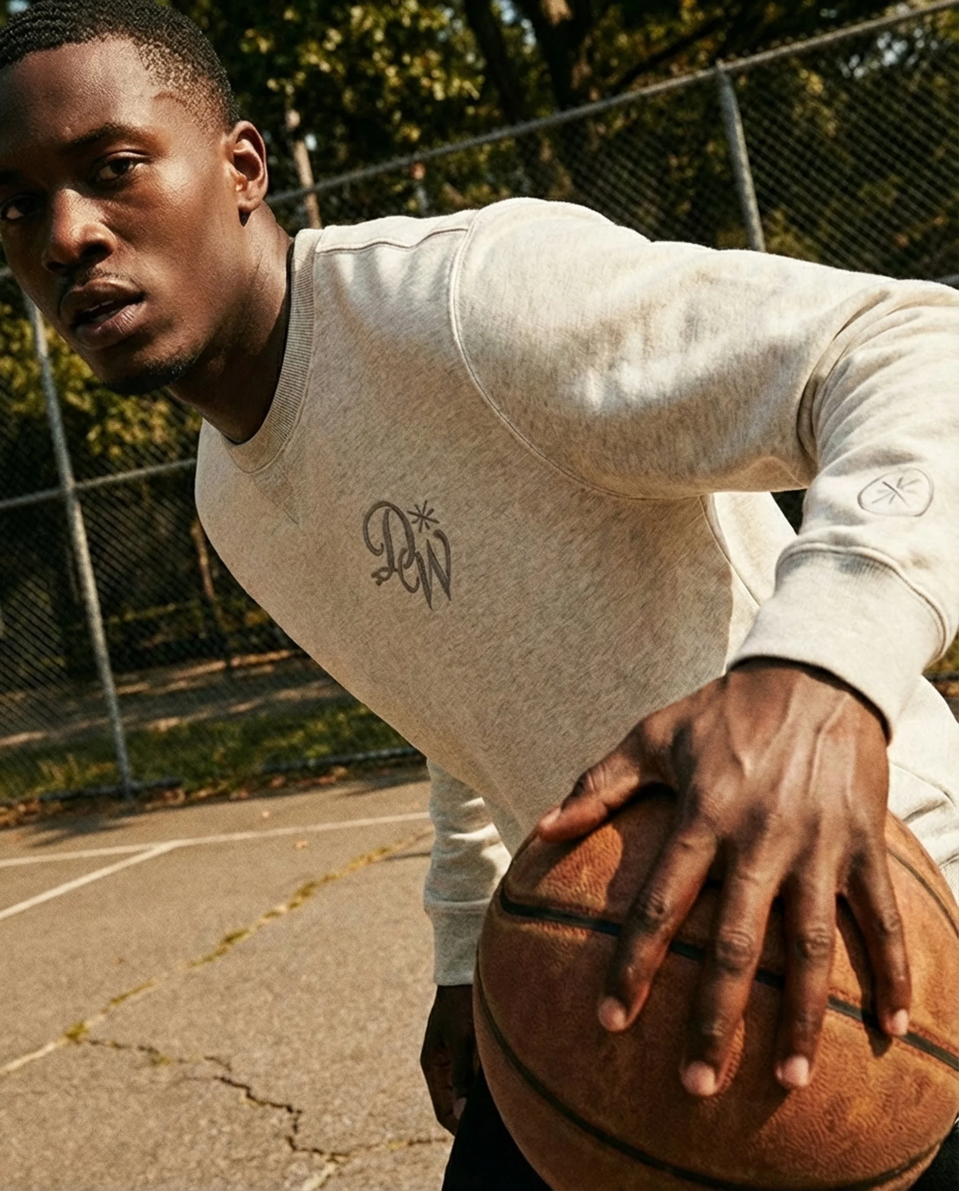 A young man in a cream-colored sweatshirt is on a basketball court at sunset, holding a basketball and looking at the camera. There is a chain-link fence and trees in the background.  AI fashion editorial.
