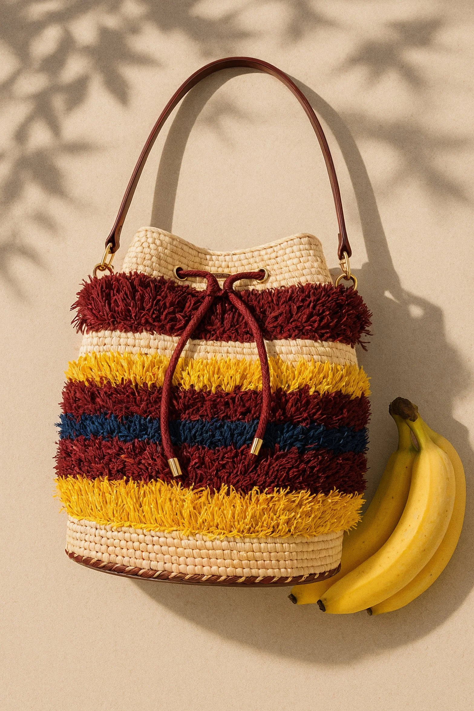 A colorful woven bag with horizontal stripes in red, yellow, blue, and beige, hanging against a plain beige background. A bunch of bananas is placed beside it on the right side of the image.