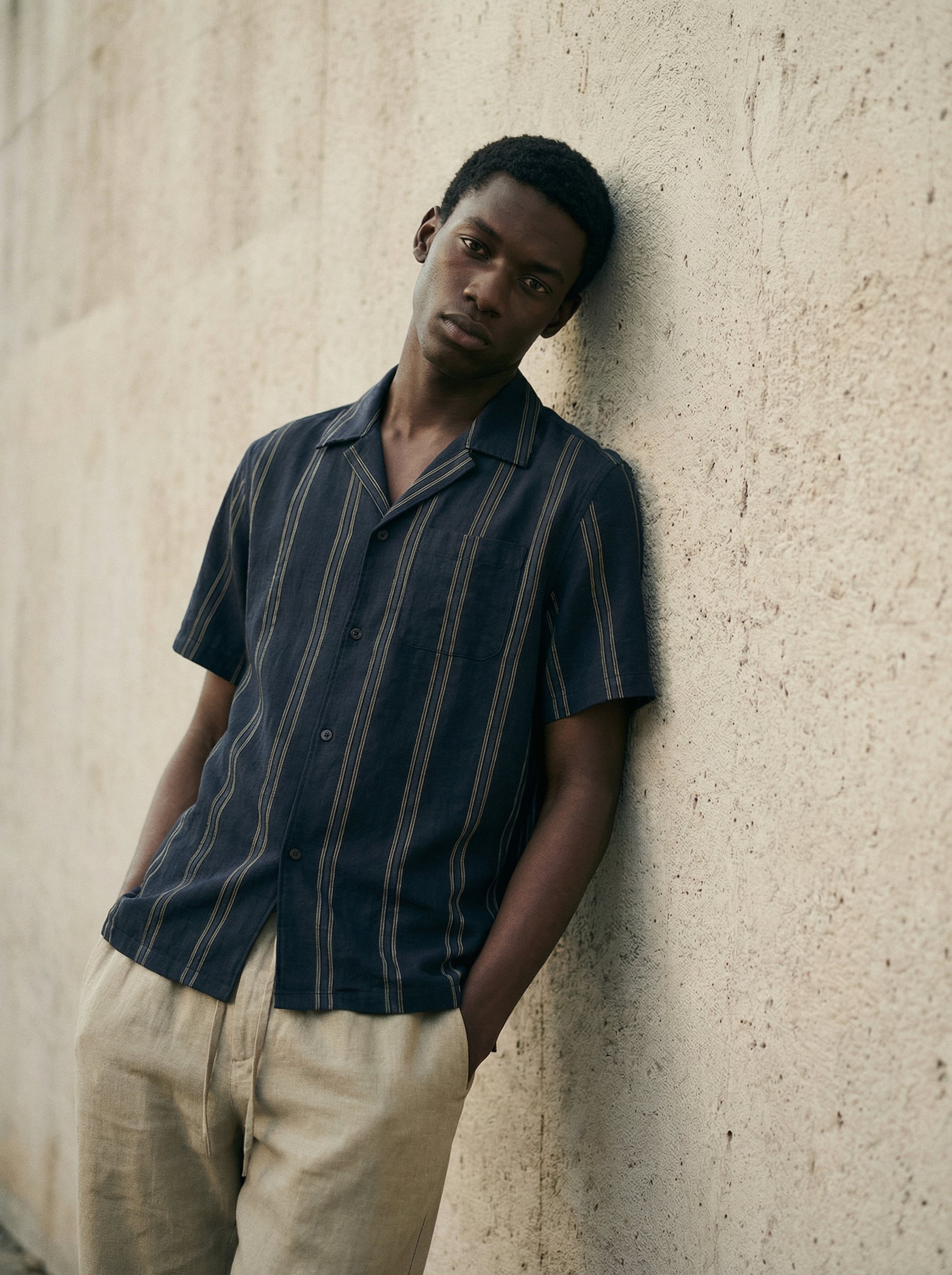 A young man with dark skin, short curly black hair, and a serious expression, leaning against a textured off-white wall.