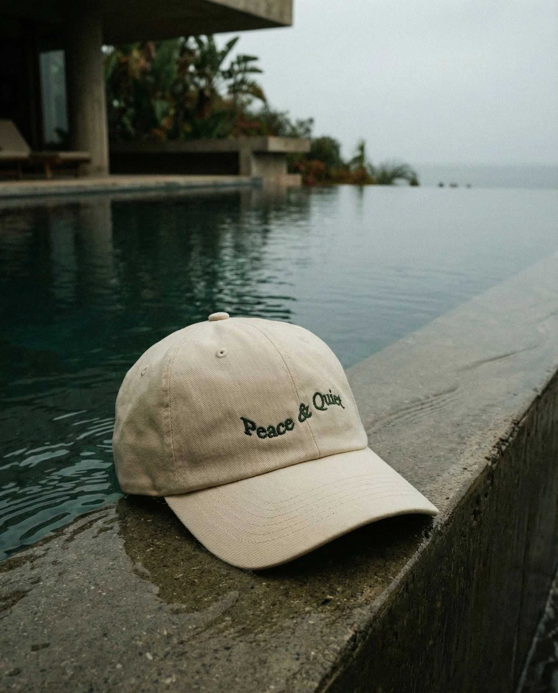 A beige baseball cap resting on the edge of a concrete poolside, with a view of the pool and greenery in the background.