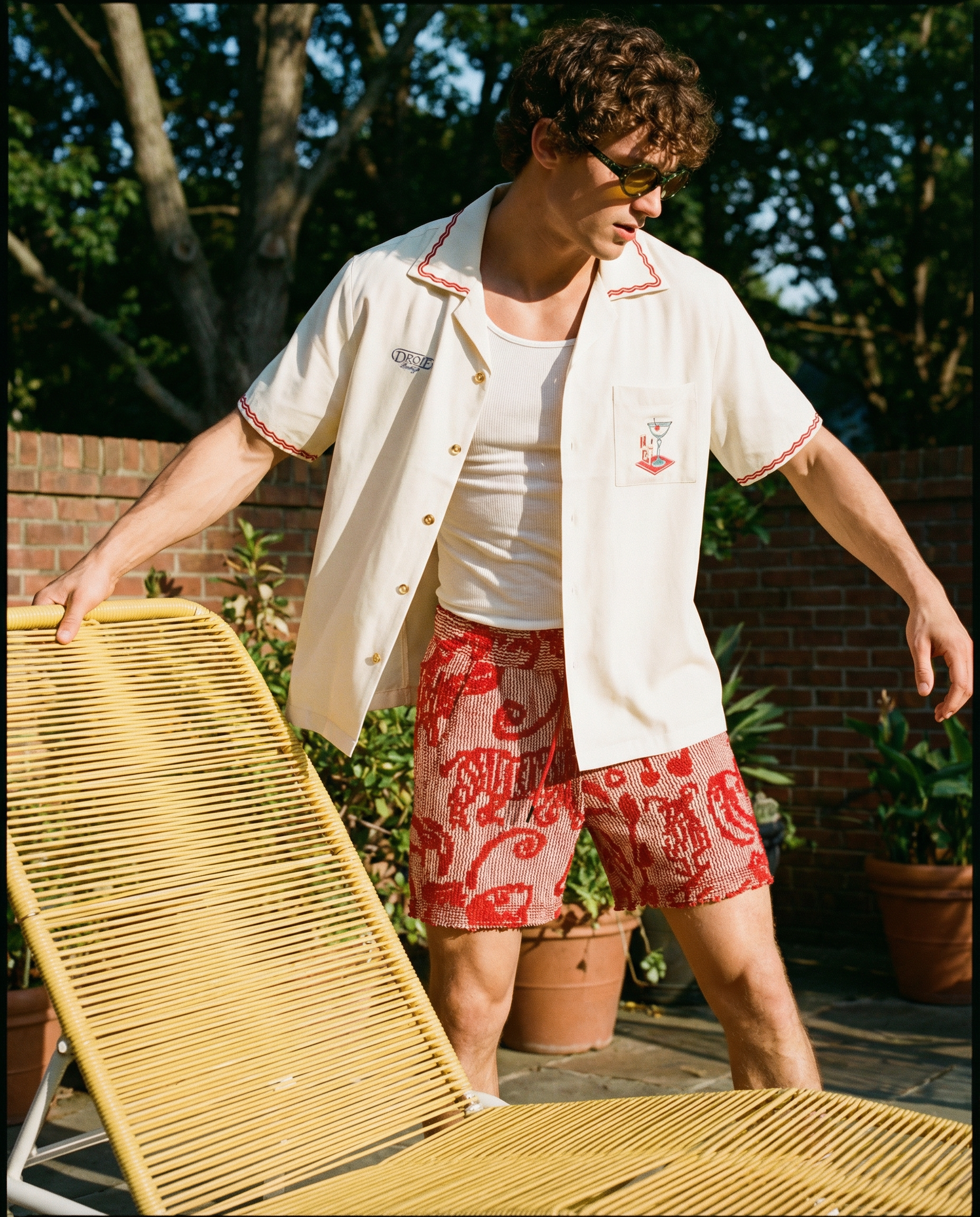 A man with curly hair and glasses is standing outdoors on a sunny day, wearing a cream-colored short-sleeve shirt with red piping, a white tank top, and red patterned shorts. He appears to be leaning with his hands on a yellow lounge chair, possibly 