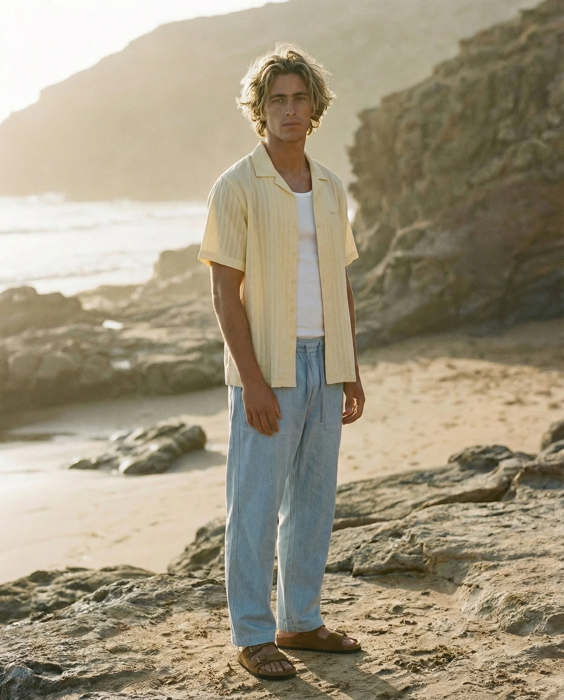 A young man stands on a sandy beach with rocky formations and cliffs in the background, wearing a yellow short-sleeved shirt over a white T-shirt, loose light blue jeans, and brown sandals, with the sun setting behind him.  AI fashion editorial image