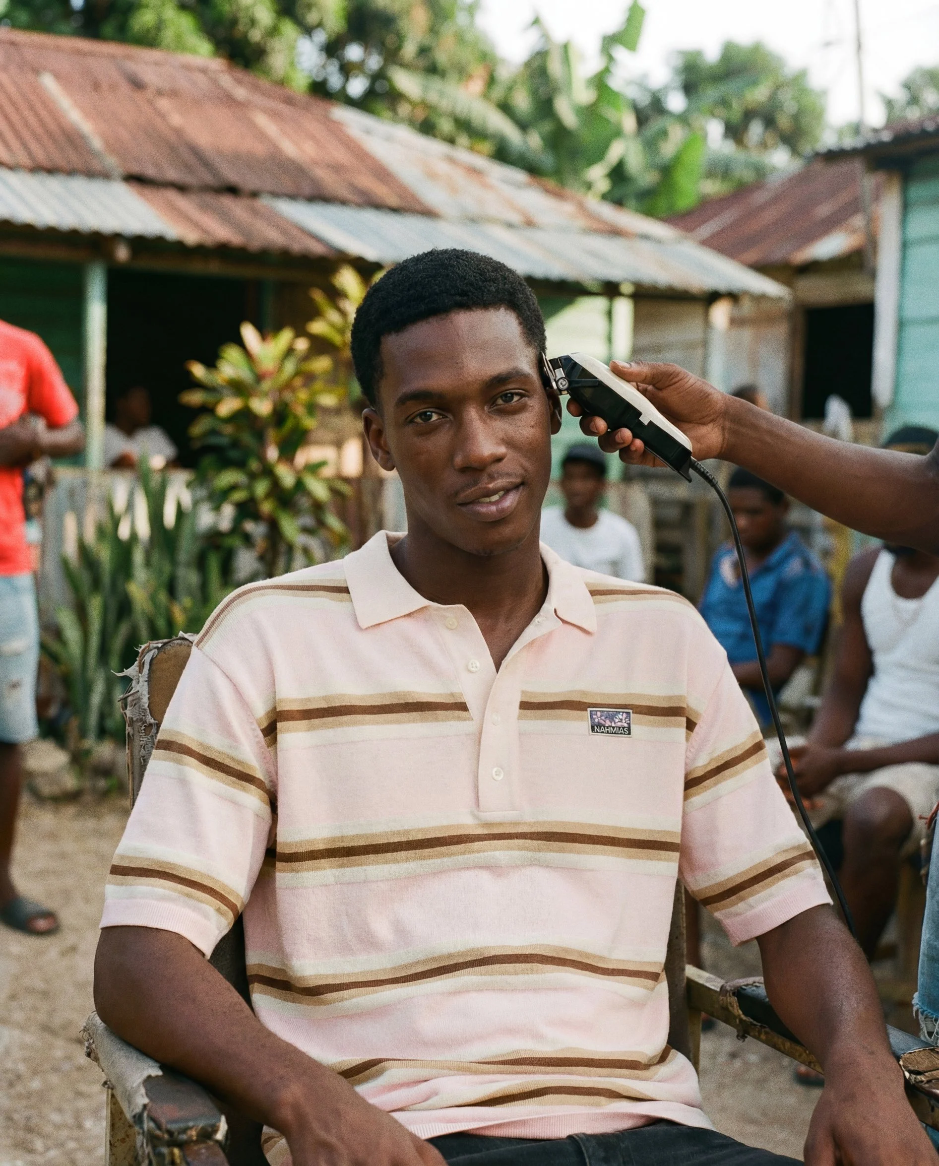 A young man with dark skin and short black curly hair getting a haircut outdoors, sitting in a chair, with a barber using hair clippers on the side of his head. People and makeshift structures with corrugated metal roofs are visible in the background