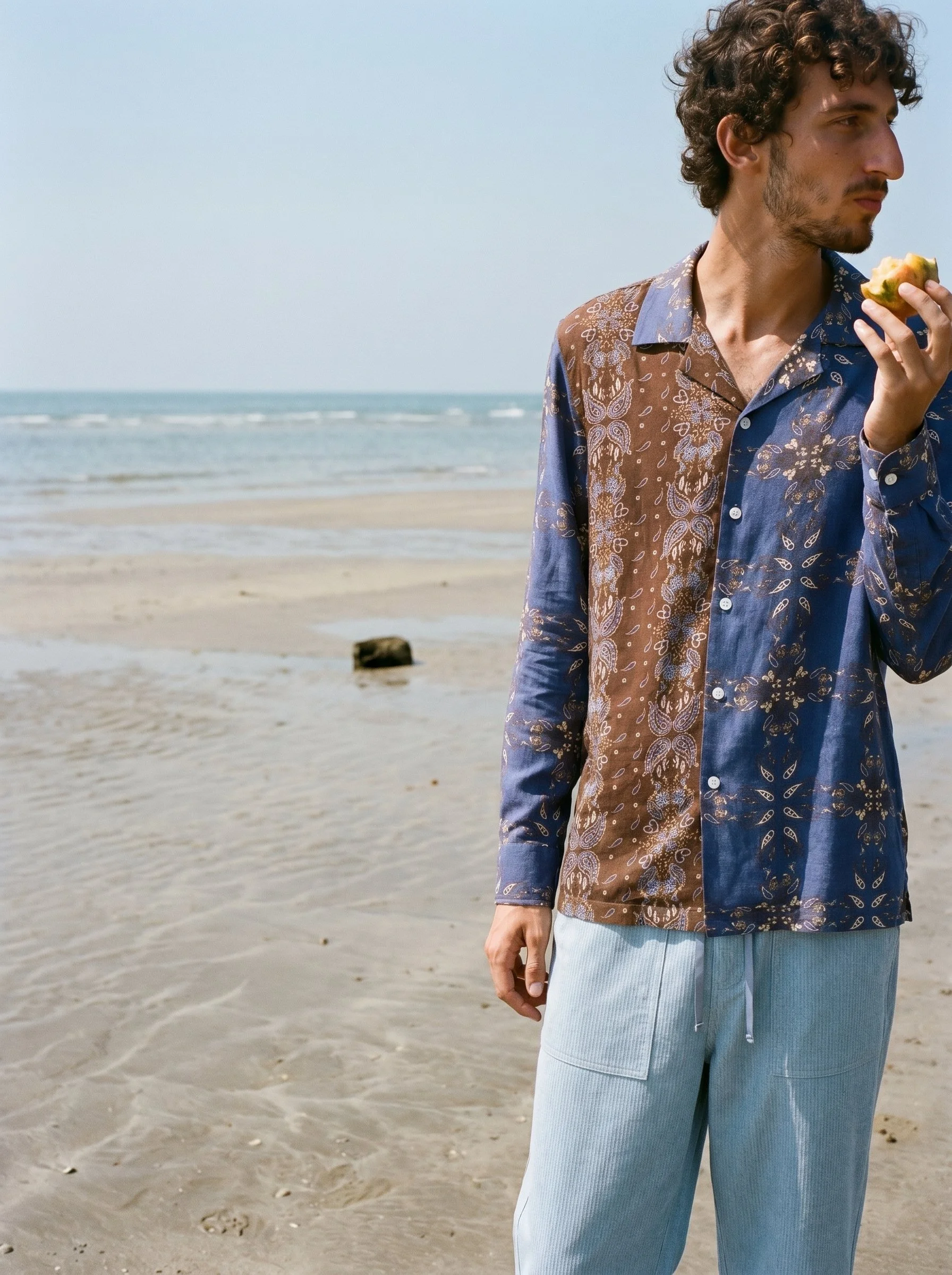 A young man with curly hair and a beard standing on a sandy beach, holding a slice of fruit, with the ocean and a light blue sky in the background.