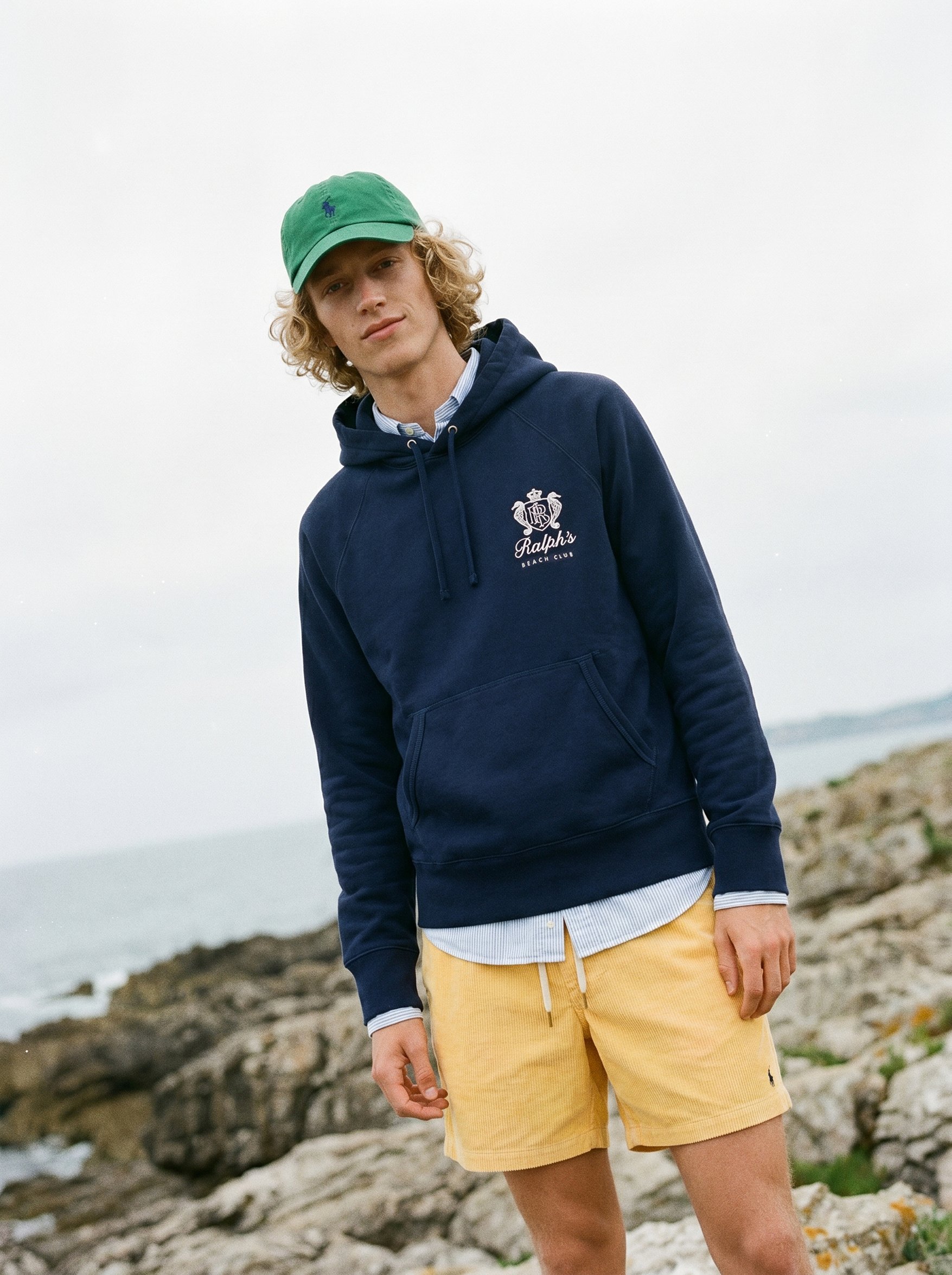 A young man with curly blonde hair wearing a green cap, navy blue hoodie with Ralph's Beach Club logo, white shirt, and yellow shorts, standing on a rocky beach shoreline with ocean waves in the background.