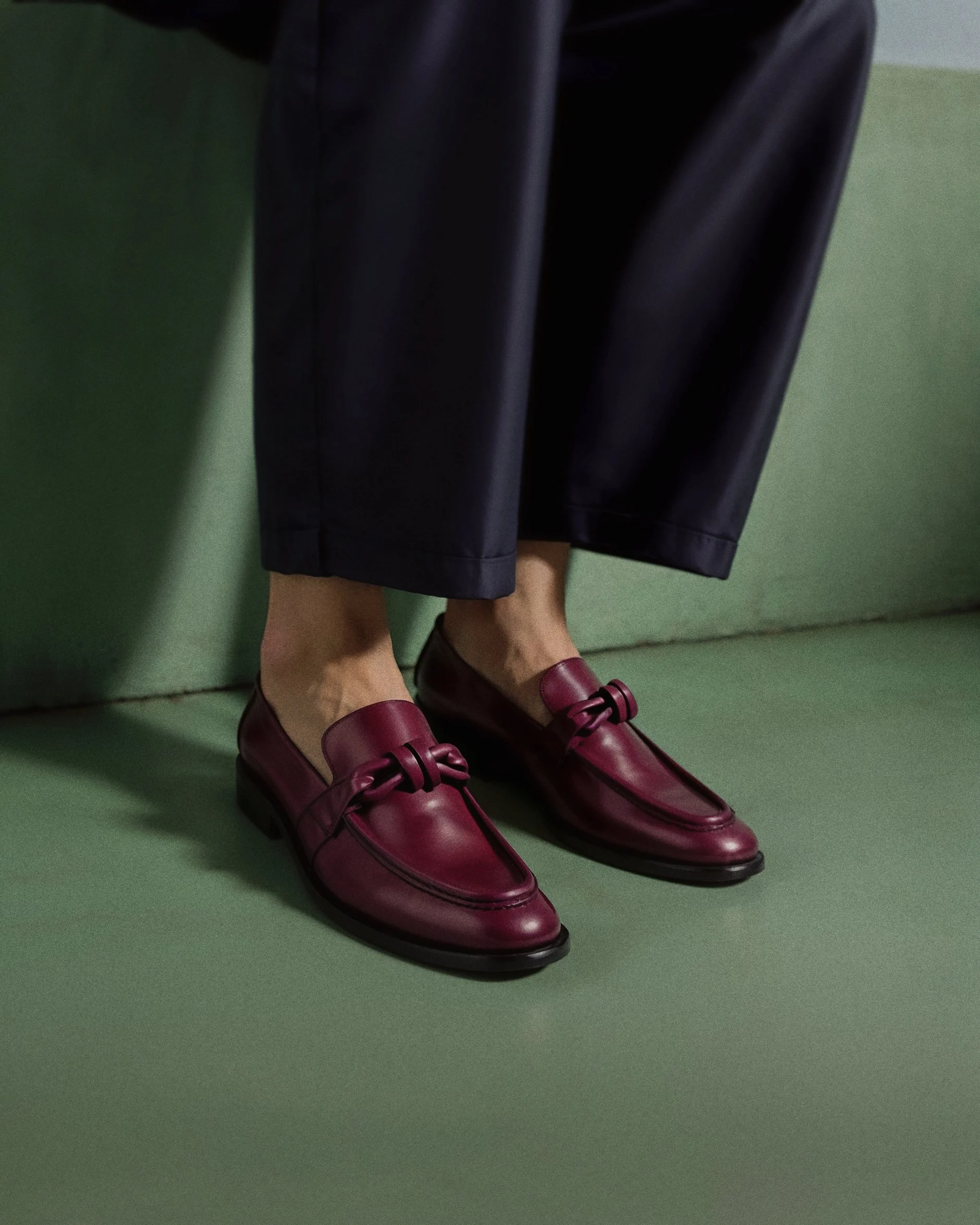 Close-up of legs wearing black leather pants and burgundy shoes with decorative bows, positioned against a green background.  AI fashion editorial.