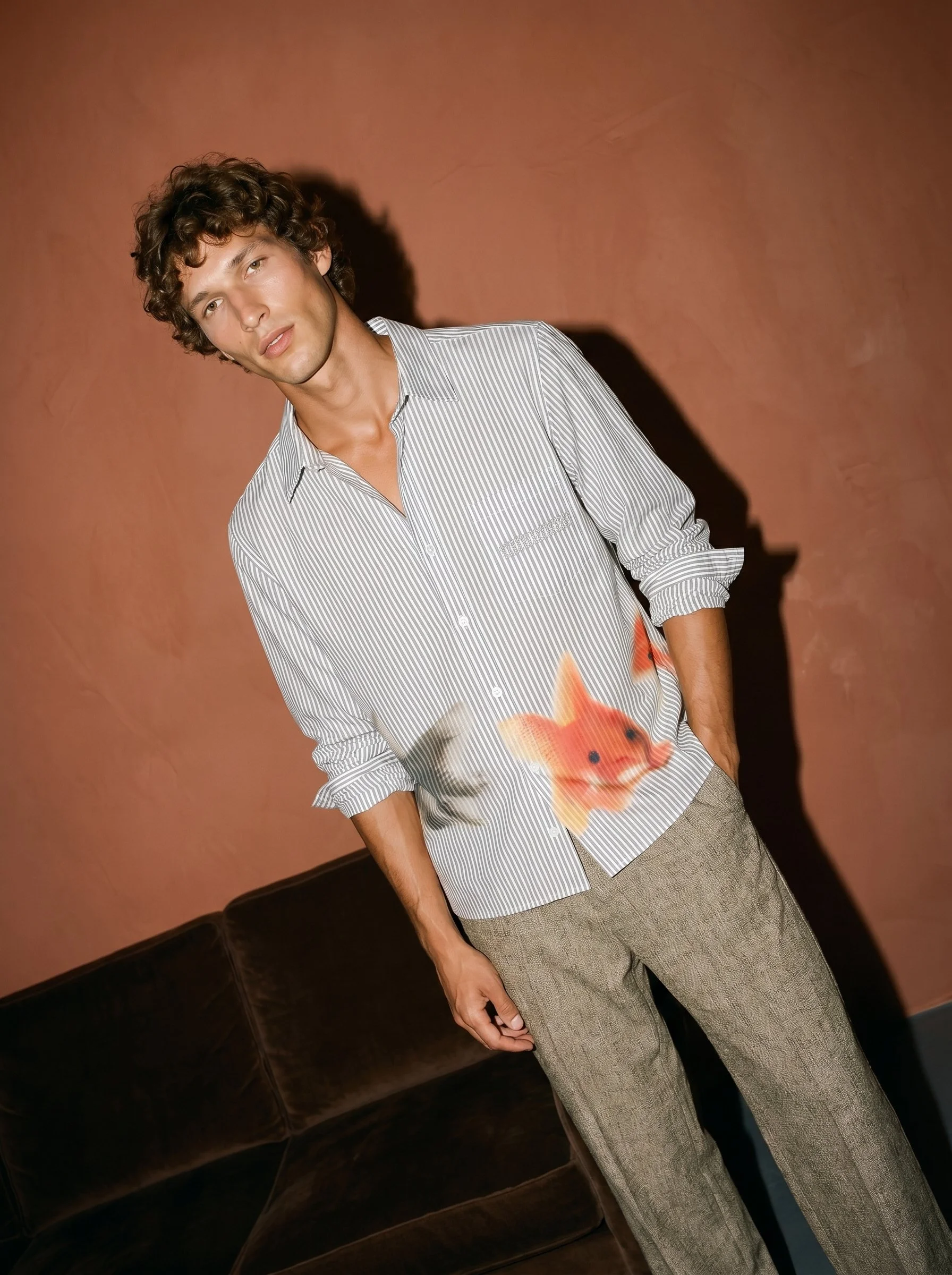 A young man with curly brown hair wearing a light blue and white striped shirt and beige plaid pants standing inside against a brown wall. There is a fish graphic on his shirt, and a dark brown couch is partially visible at the bottom left corner.