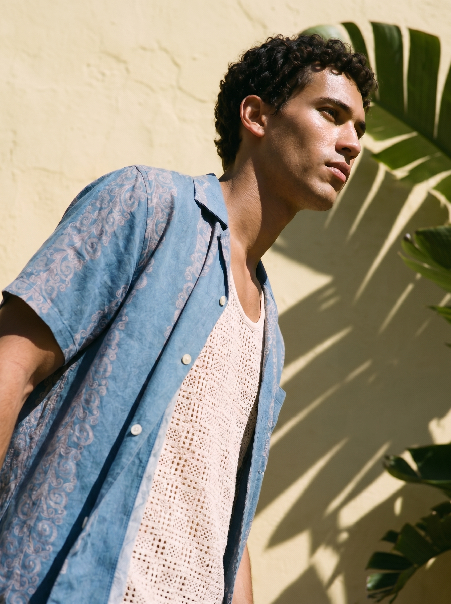 A person with short curly dark hair and light skin, wearing a white crochet tank top and a blue patterned shirt, standing outdoors near a yellow wall with shadows of large green leaves.