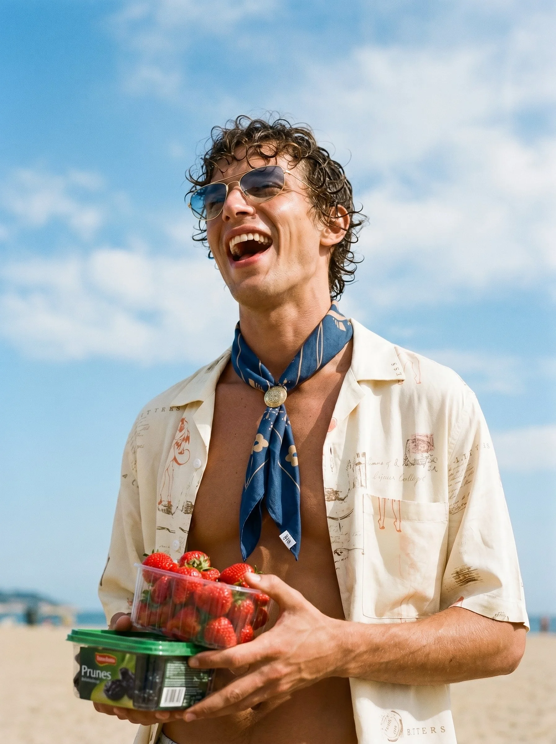 A man with curly hair, wearing sunglasses and a patterned shirt, stands outdoors on a sunny day, holding a container of strawberries and a smaller container of prunes.