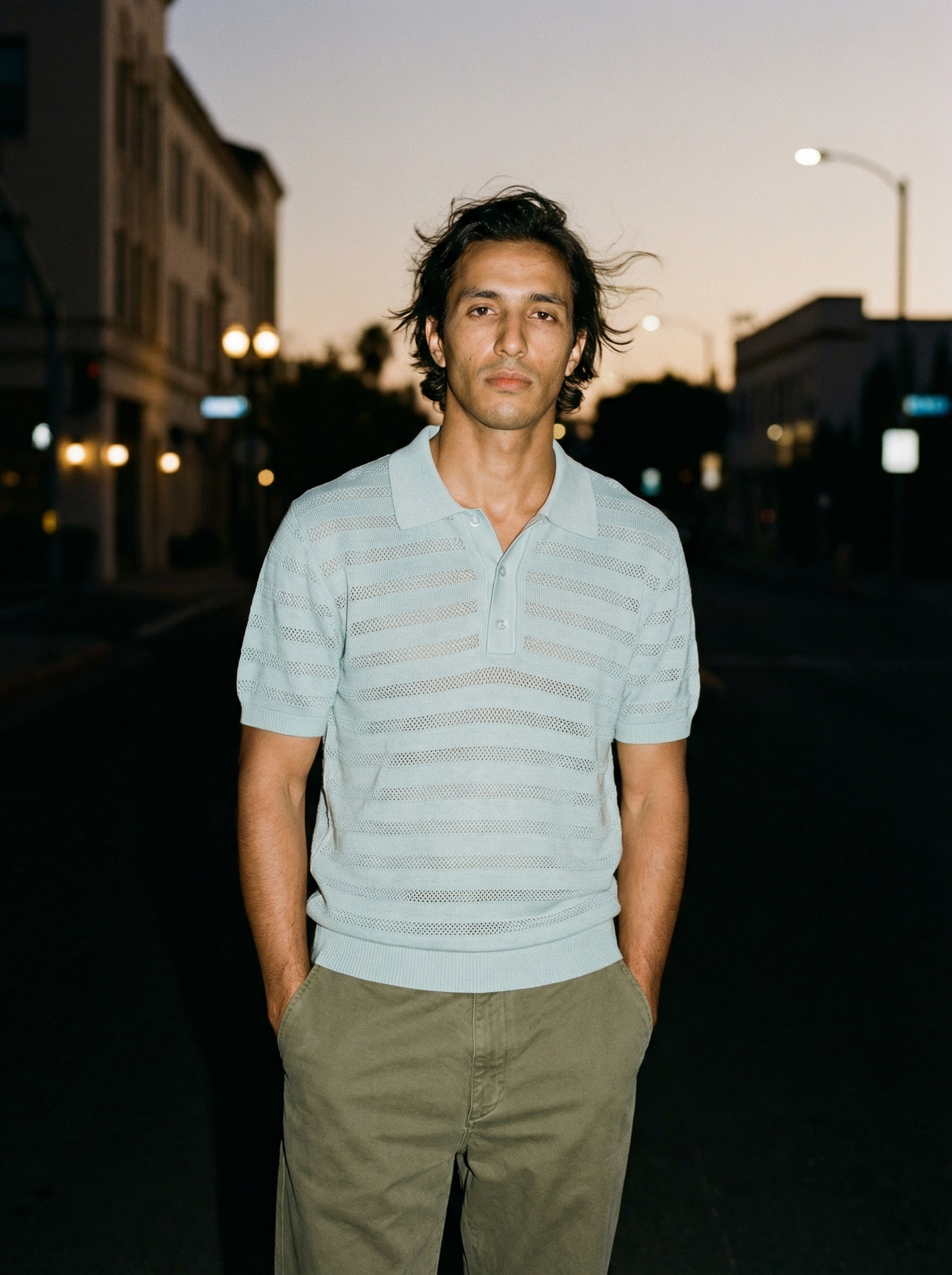 Young man with dark hair standing outdoors during dusk, wearing a light blue polo shirt and khaki pants.