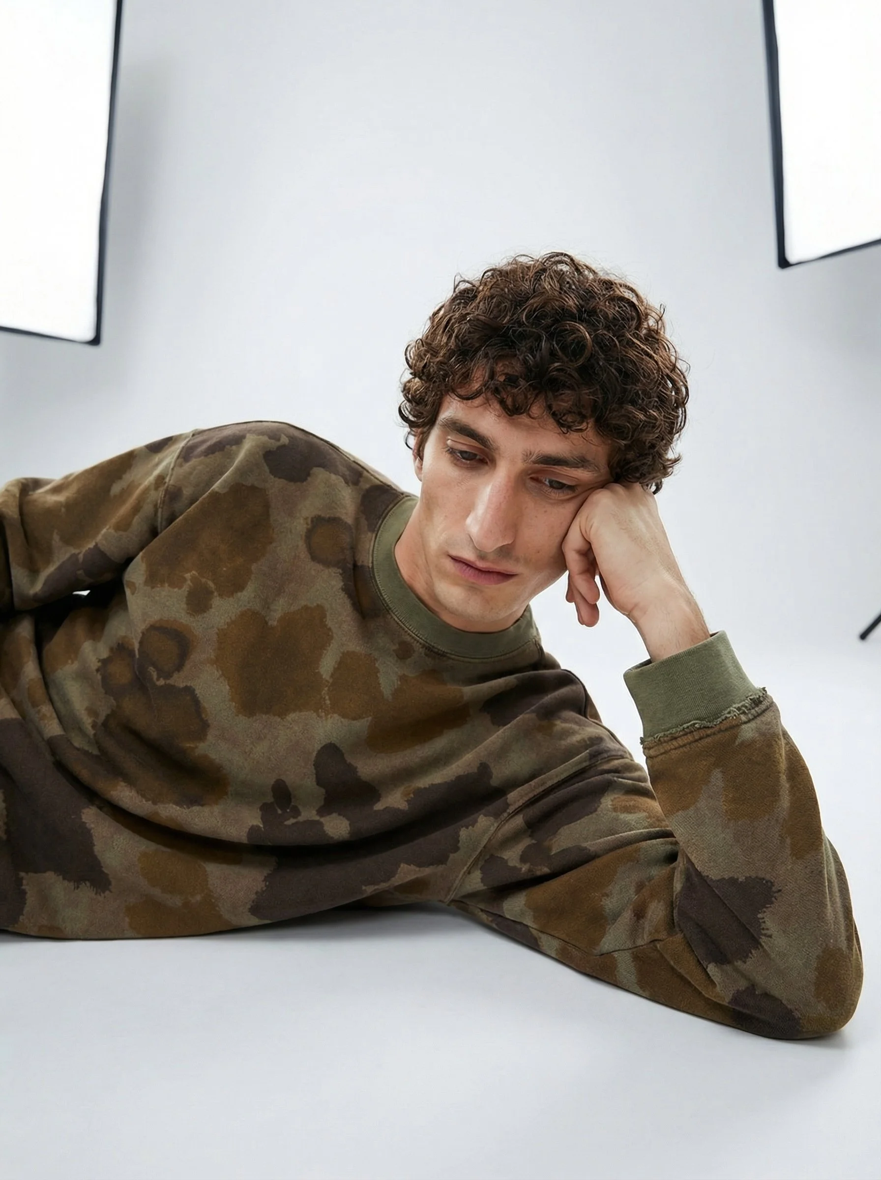Young man with curly hair wearing a camouflage sweatshirt, resting his head on his hand, in a studio with a white background and large softbox lights.