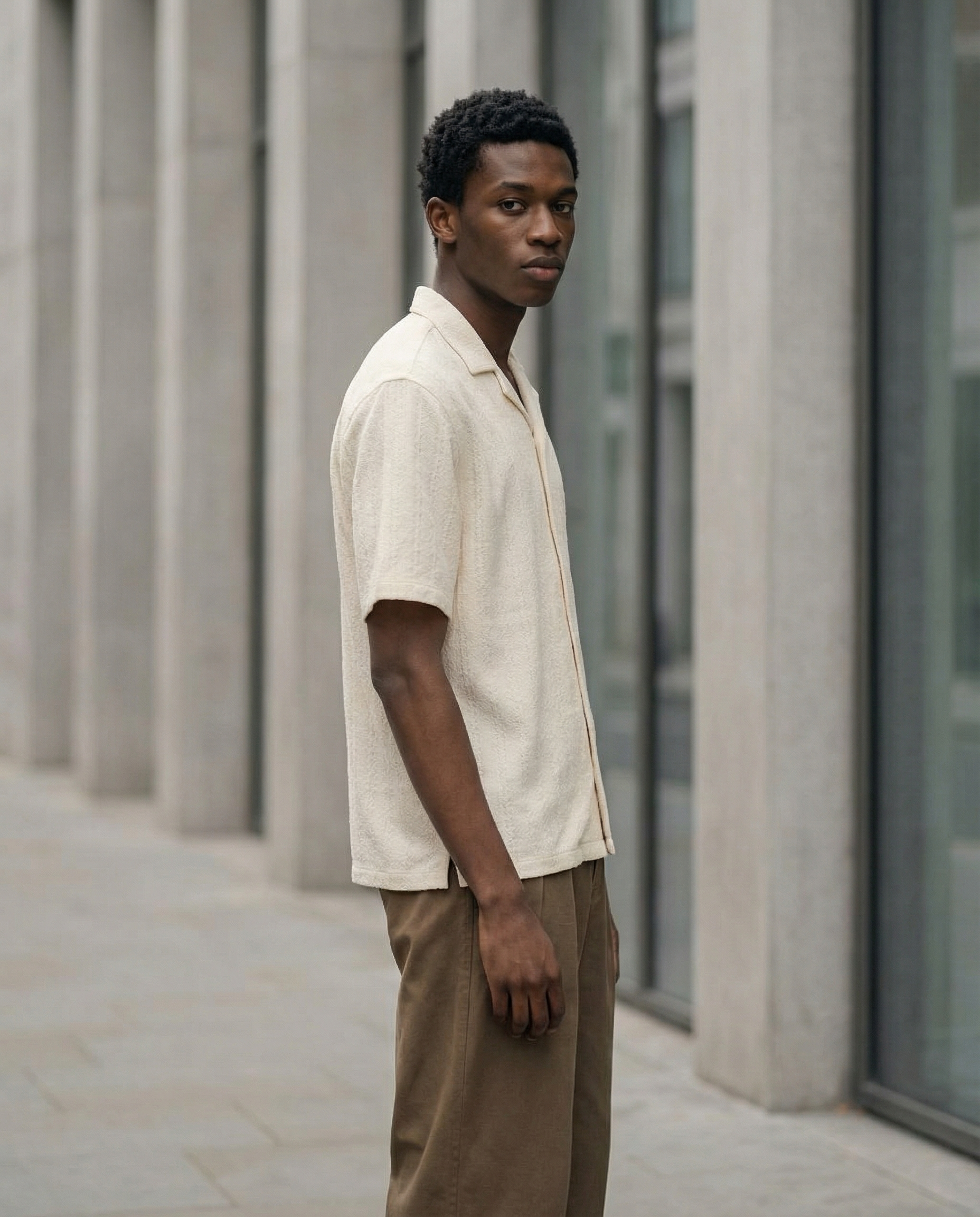 A young black man with short, curly hair stands outdoors against a gray building with large glass windows, wearing a beige shirt and brown pants, looking directly at the camera with a serious expression.  AI fashion editorial.