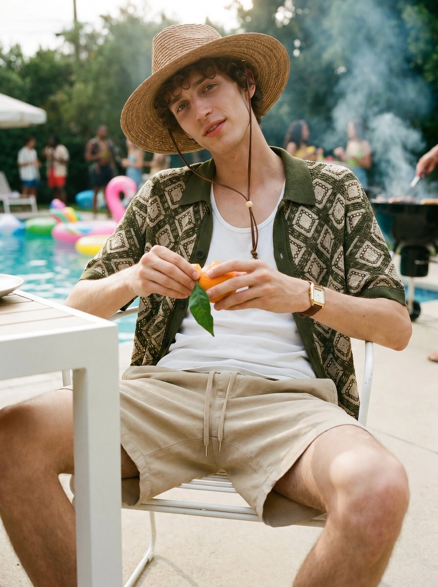 A young man sitting outdoors at a gathering near a swimming pool, wearing a wide-brimmed straw hat, a patterned shirt, and beige shorts, with indoor furniture and other people in the background.