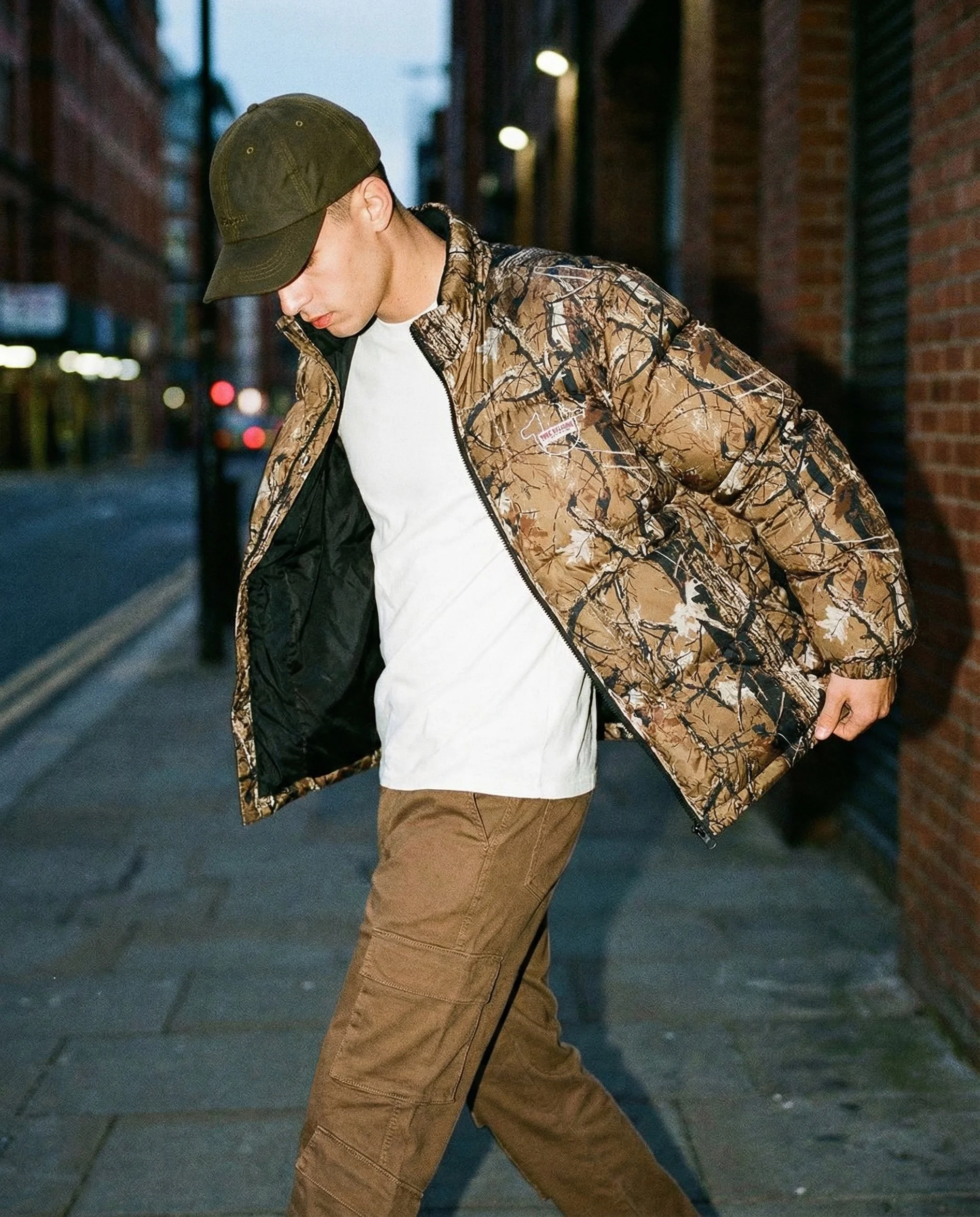 A young man wearing a camouflage jacket, white t-shirt, brown pants, and a black baseball cap walking along a city street at dusk.