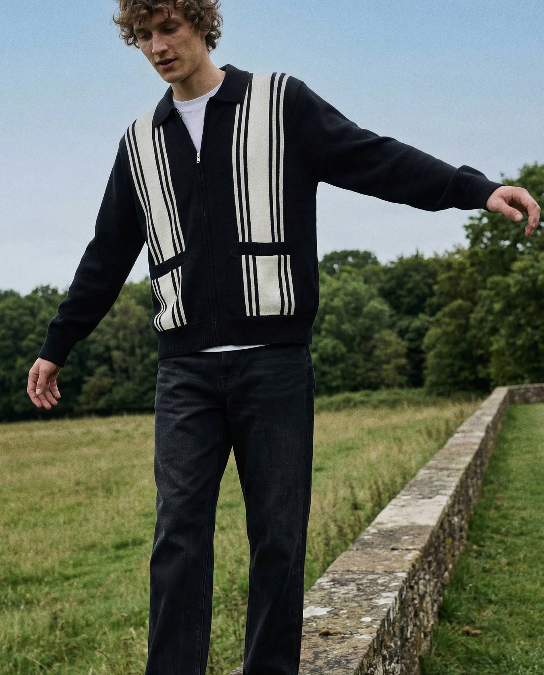 Young man in a black jacket with white and black vertical stripe design, standing outdoors on a grassy area next to a stone wall, looking down with his arms slightly outstretched.