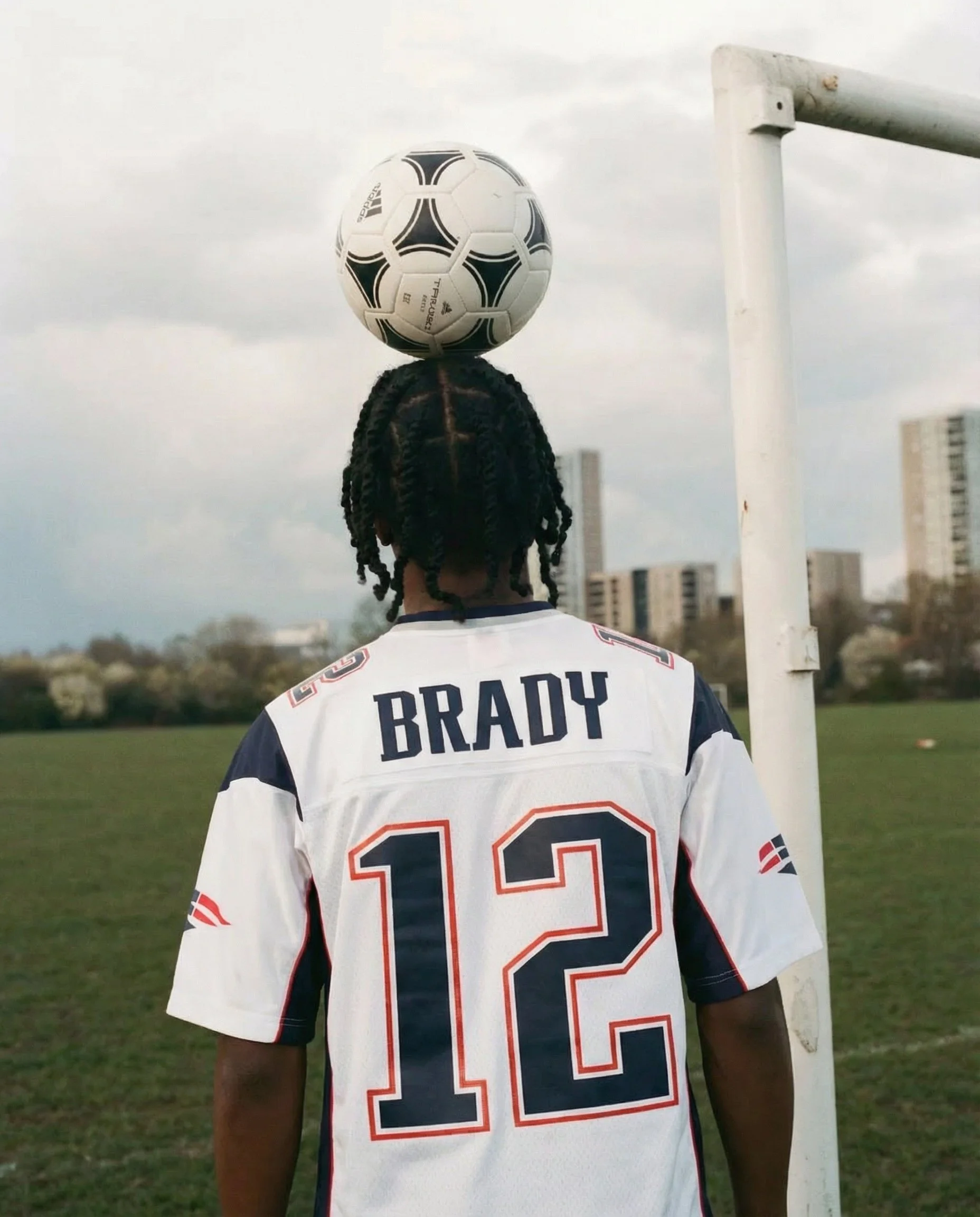 Back view of a football player wearing a white jersey with the name 'BRADY' and the number 12, standing on a football field near a goalpost, with city buildings in the background, during daytime.