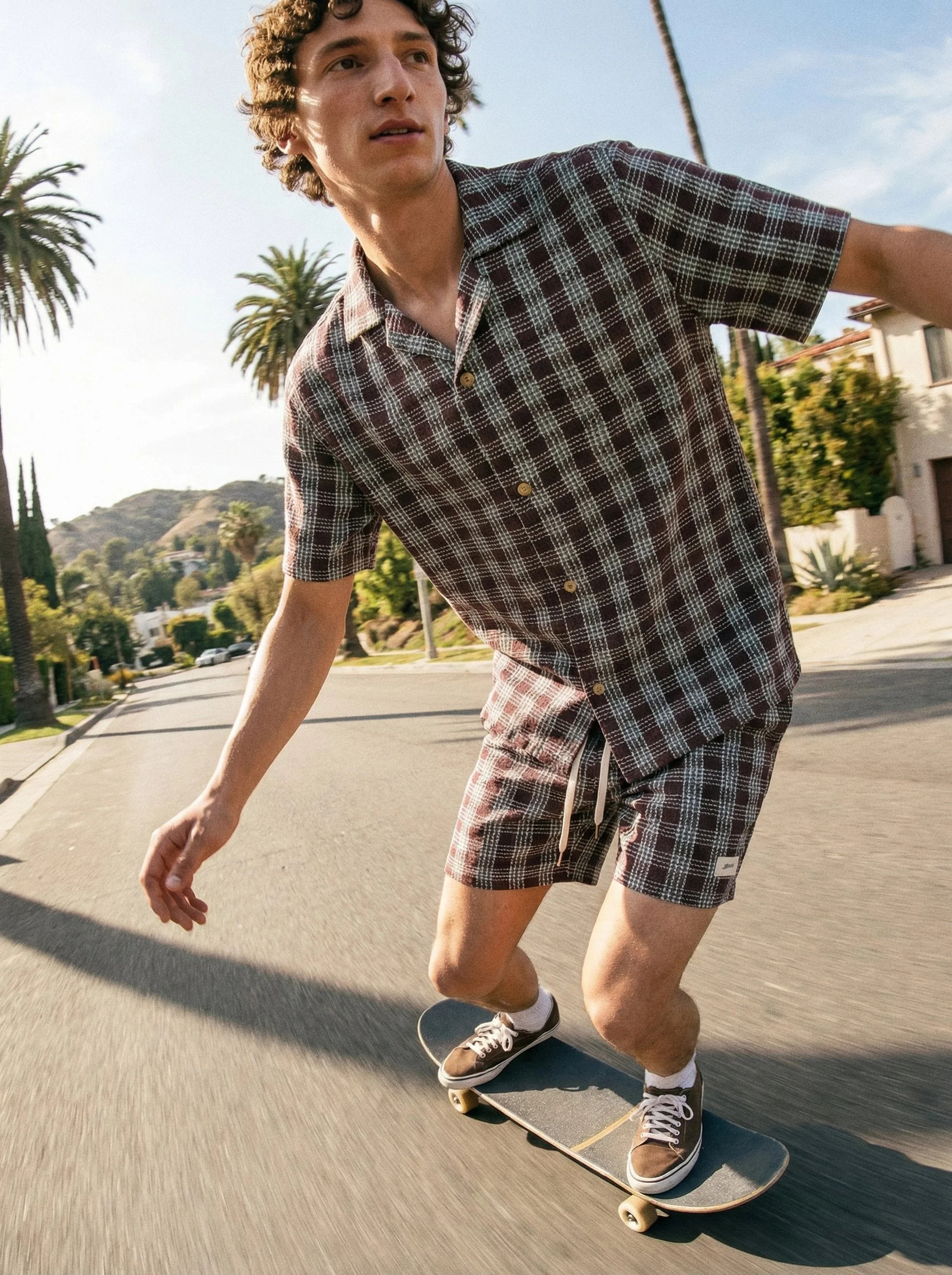 A young man with curly hair skateboarding on a sunny street, wearing a checkered short-sleeve shirt, matching shorts, and brown shoes.