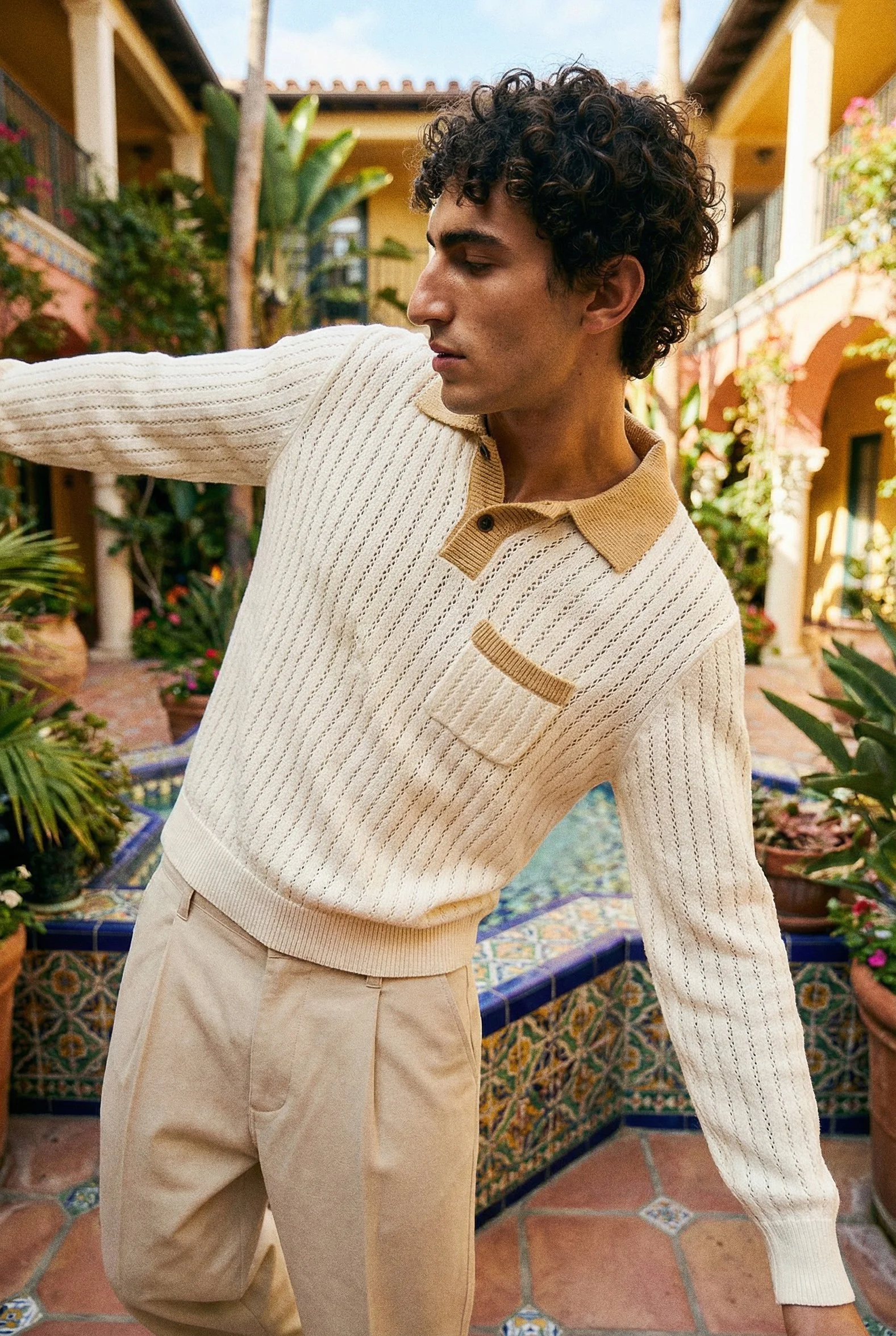 A young man with curly dark hair wearing a cream-colored sweater with a tan collar and beige pants standing outdoors in a courtyard with plants and colorful tiles.
