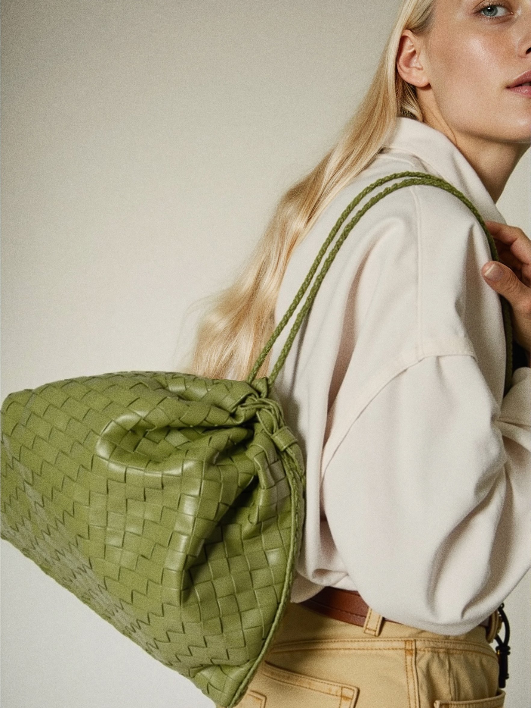 A woman in a white blazer and beige pants carrying a green woven handbag over her shoulder.