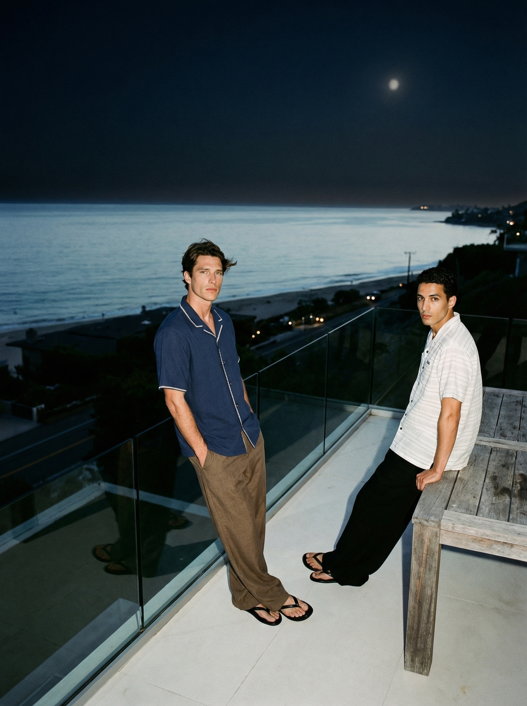 Two young men standing on a balcony at night with a view of the ocean and the moon in the sky. One wears a blue shirt and brown pants, the other a white striped shirt and black pants.