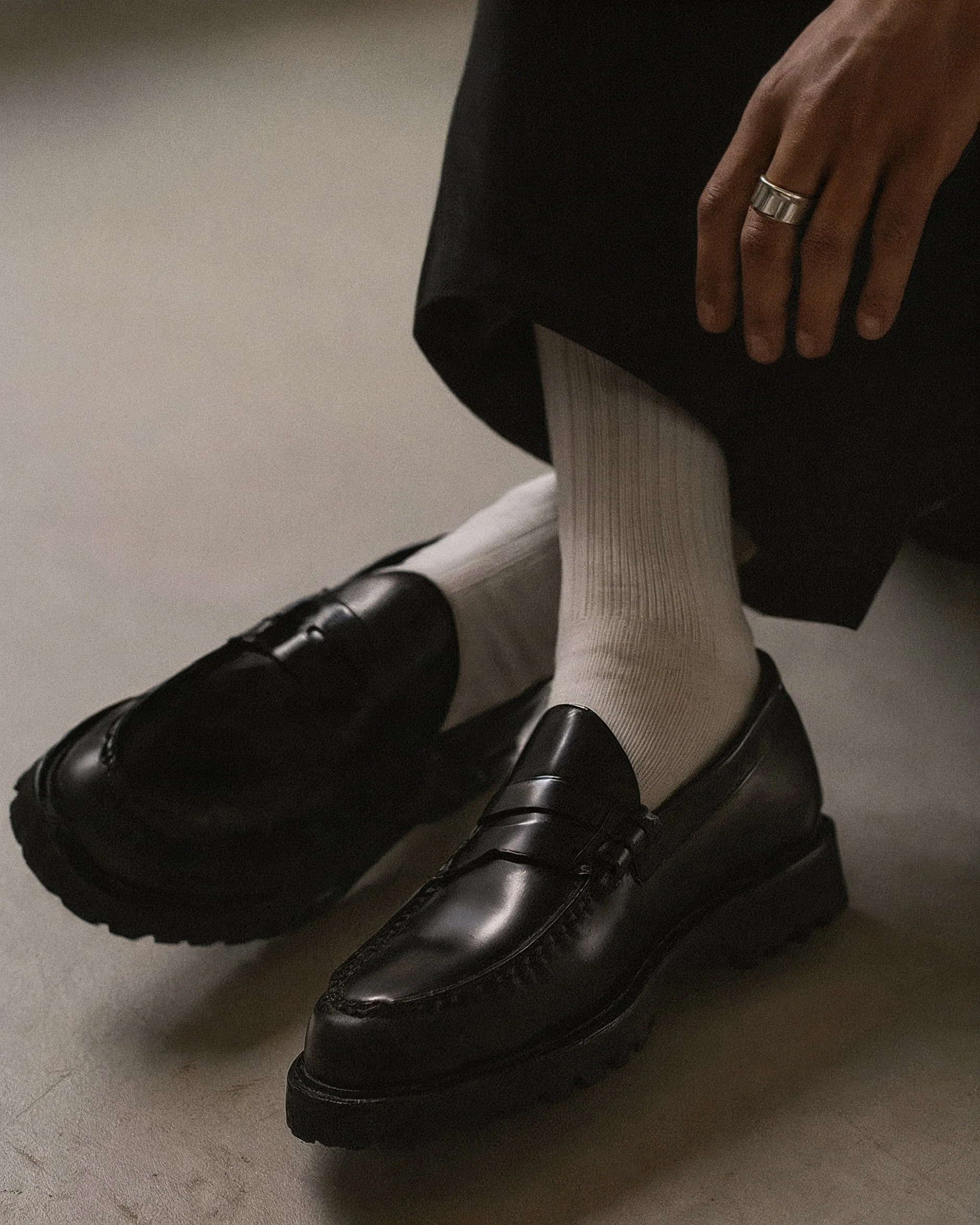 close-up of a person's leg wearing a black formal shoe, light-colored knee-high socks, and black pants, sitting on a gray floor. AI fashion editorial.