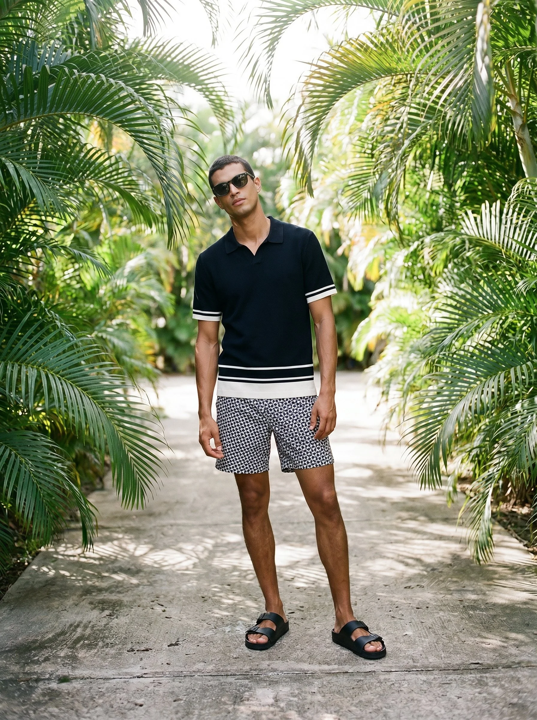 Young man wearing sunglasses, a black polo shirt with white stripes on the sleeves, patterned shorts, and sandals, standing on a pathway surrounded by lush green tropical plants.