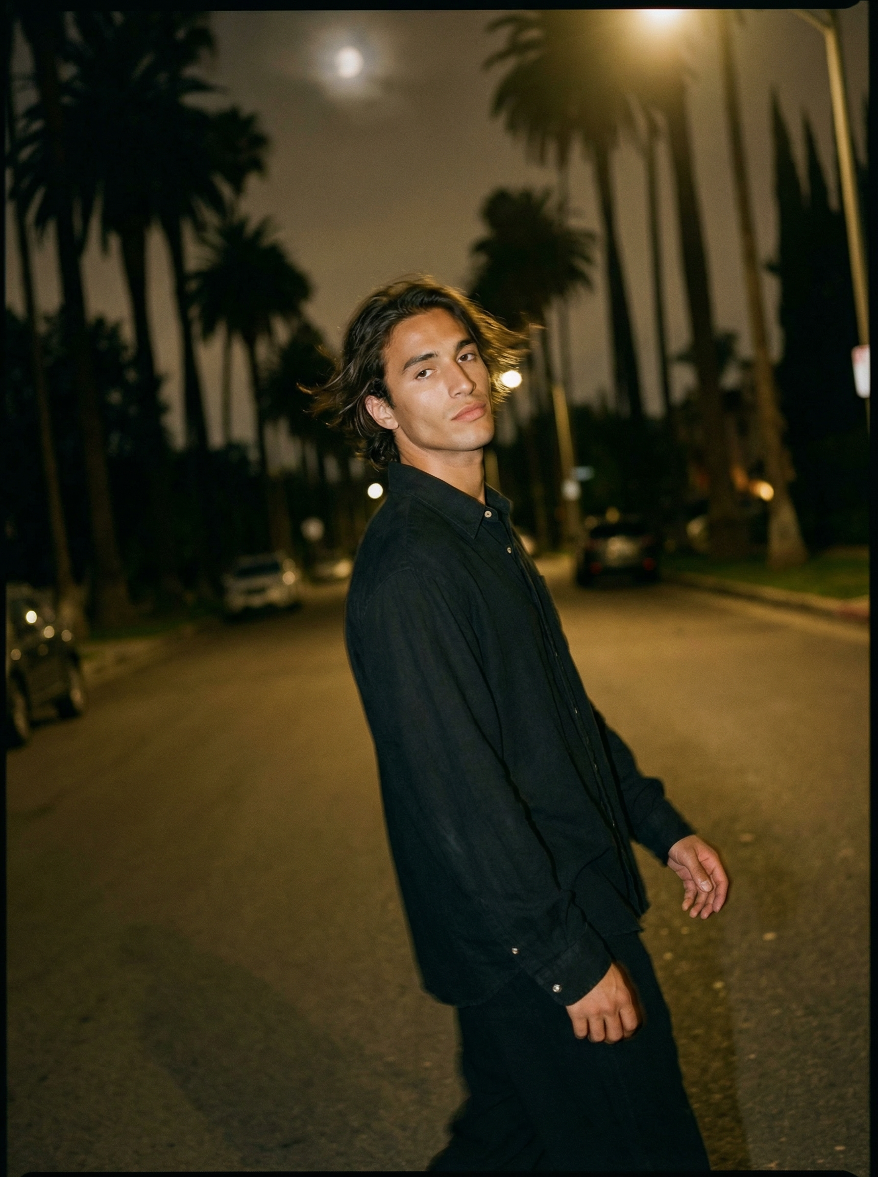 A young man standing on a street at night with palm trees and parked cars in the background under a cloudy moonlit sky.