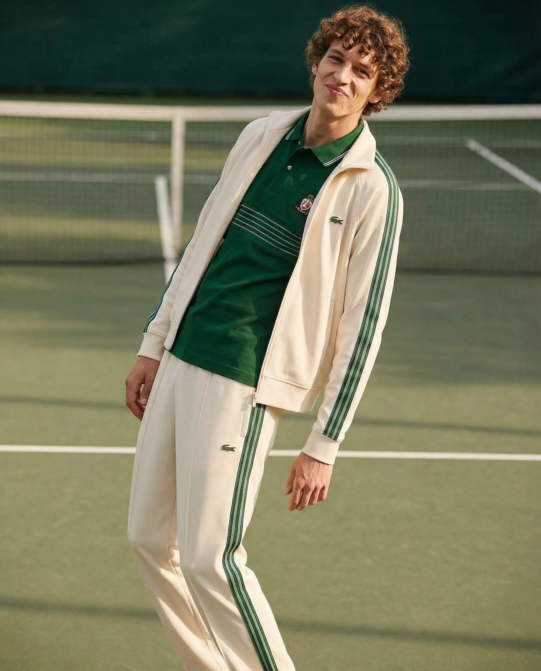 A young man with curly hair standing on a tennis court, wearing a cream-colored jacket and green Lacoste hoodie, with a smile, during daytime.