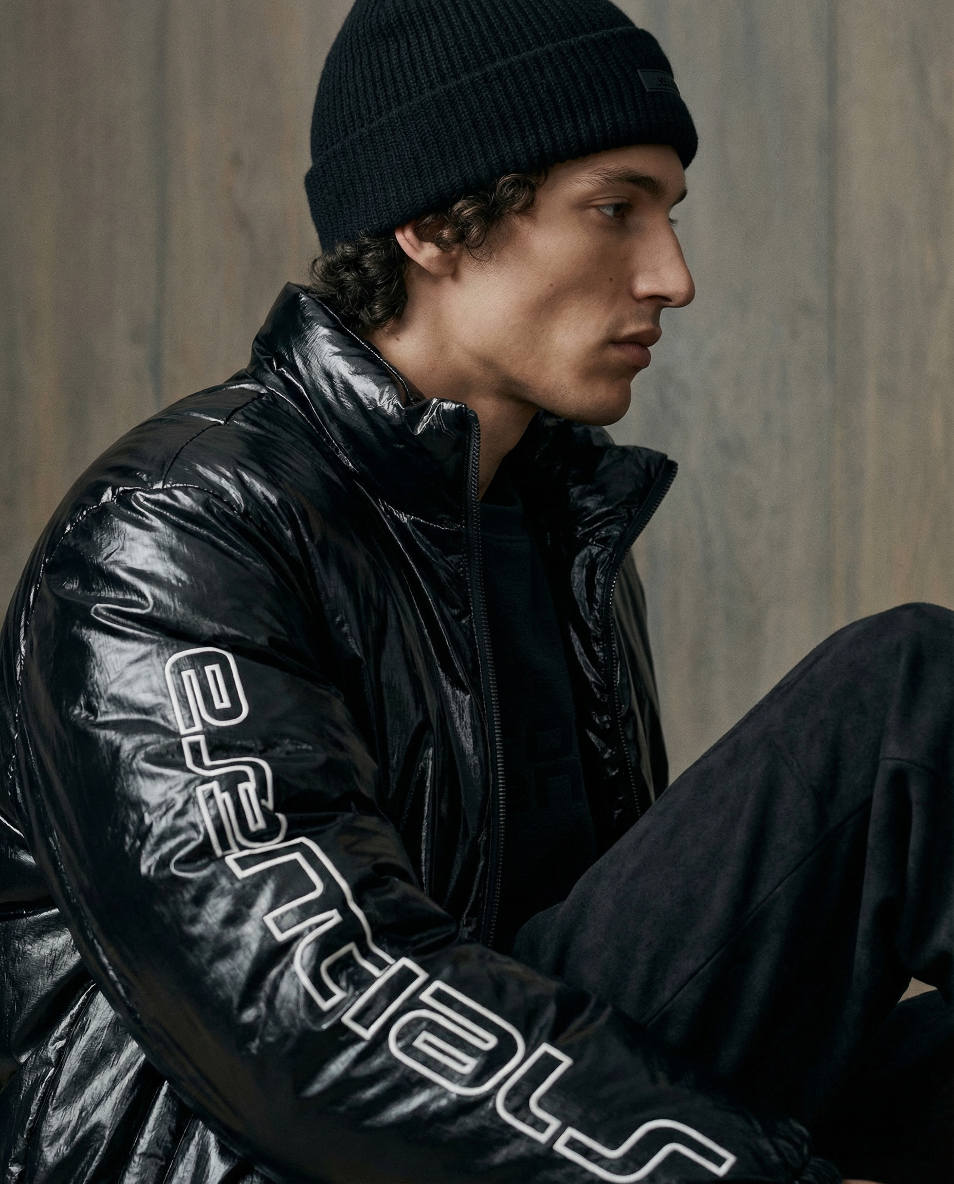 Close-up of a young man with curly hair wearing a black knit beanie and a glossy black puffer jacket, sitting against a plain wall.