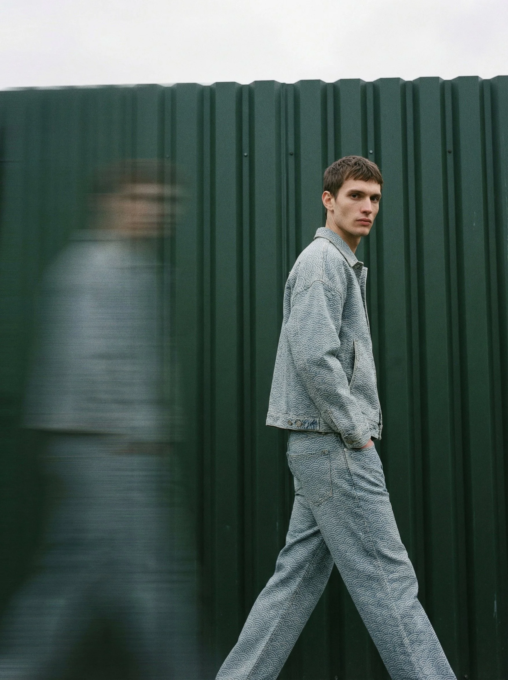 A young man with short brown hair wearing a matching patterned gray jacket and pants, walking beside a green corrugated metal fence.