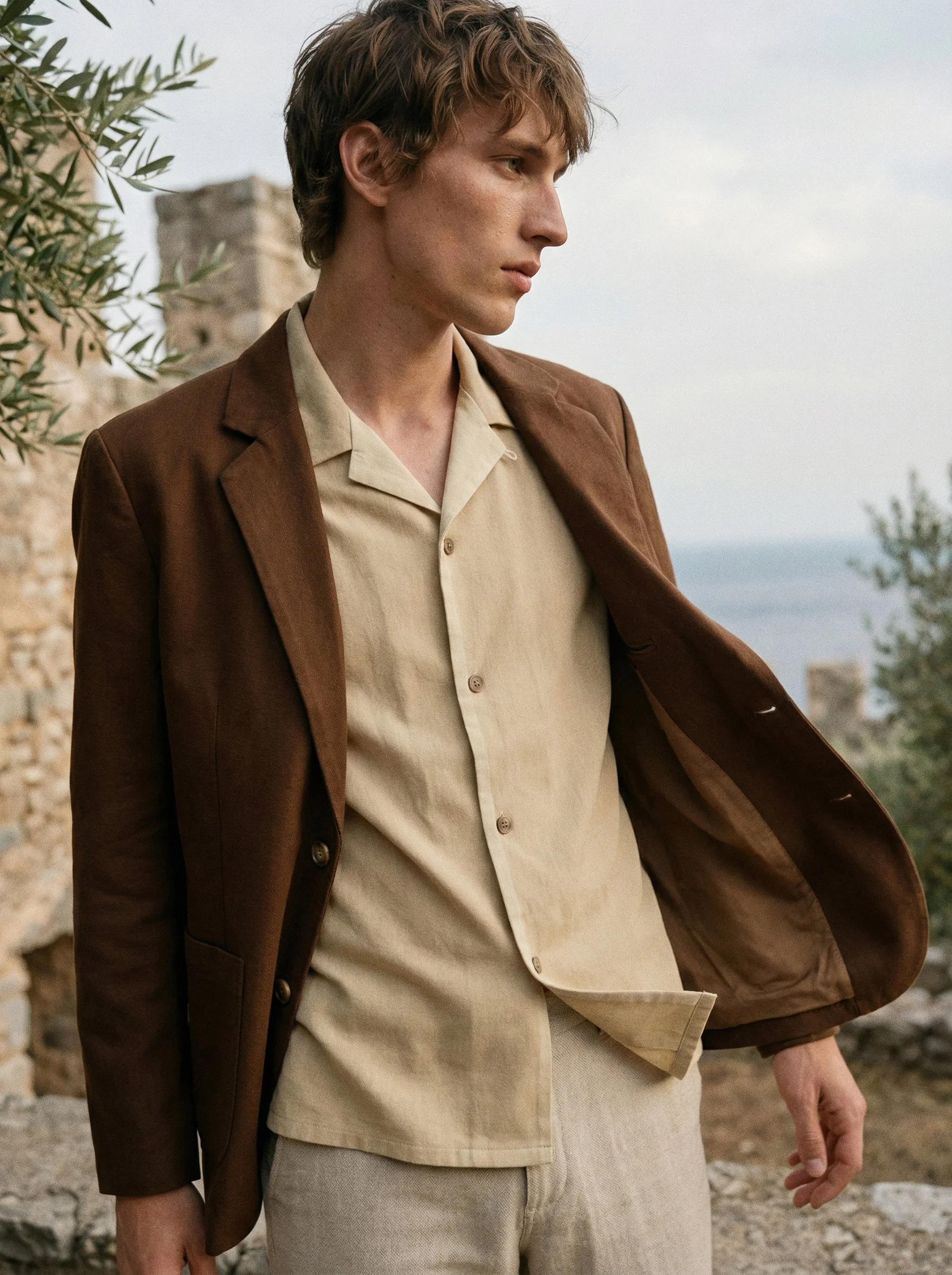 A young man with light skin and light brown hair stands outdoors, wearing a beige shirt and a brown jacket. The background features a stone structure and some greenery, with cloudy skies.