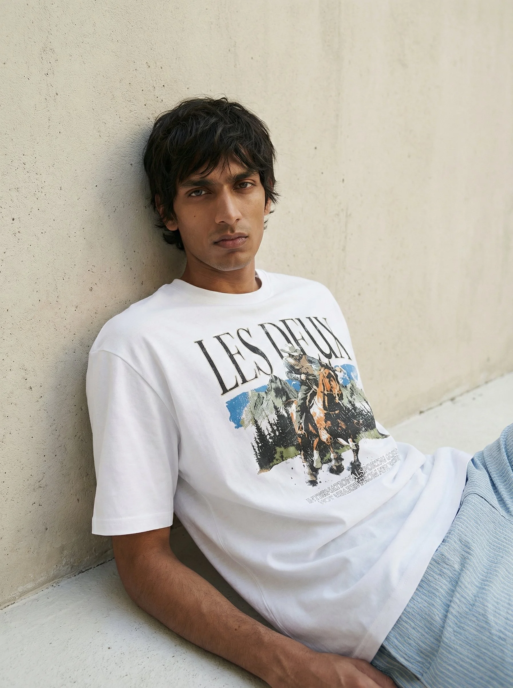Young man with dark hair, wearing a white graphic T-shirt and light-colored striped pants, leaning against a beige stucco wall.