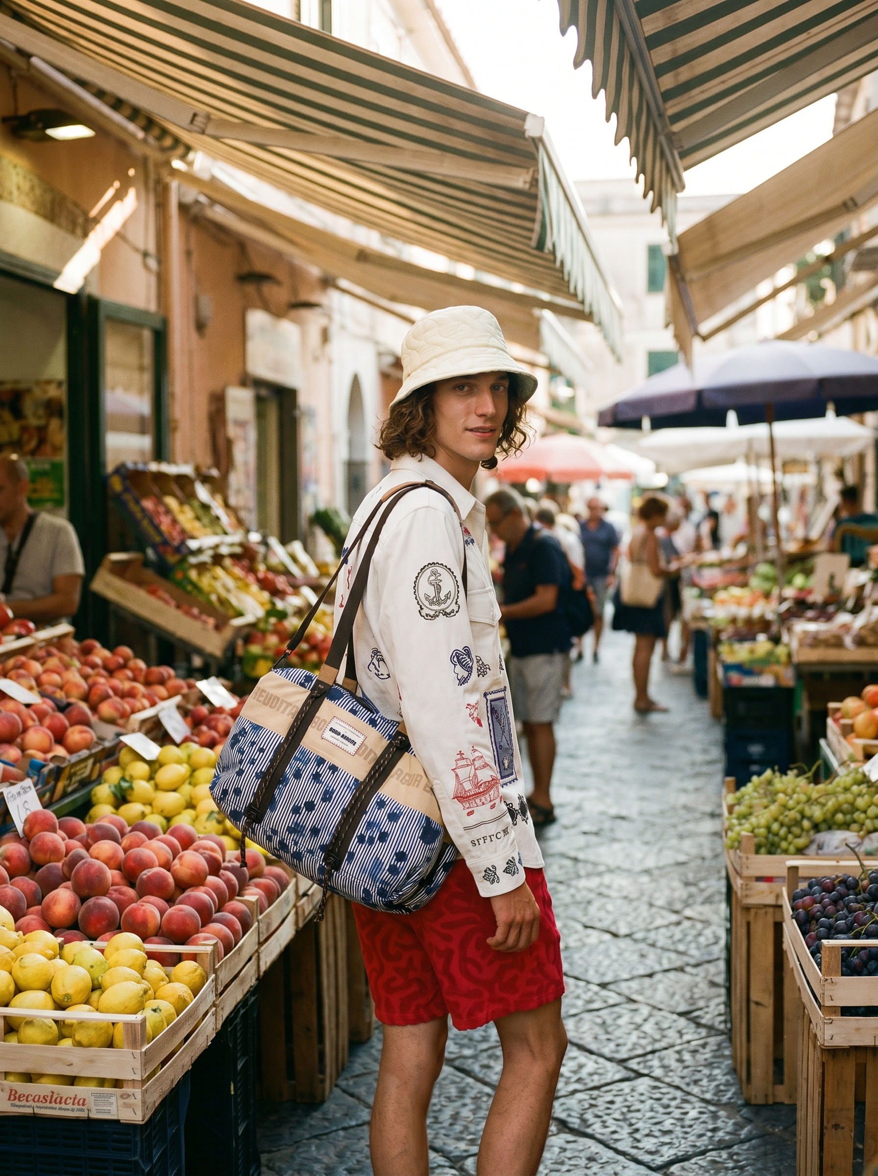 A young man with curly hair, wearing a white bucket hat and a white long-sleeve shirt with embroidered patches, stands at an outdoor fruit market. He carries a large striped tote bag. The market is shaded with striped awnings, and there are fruits li