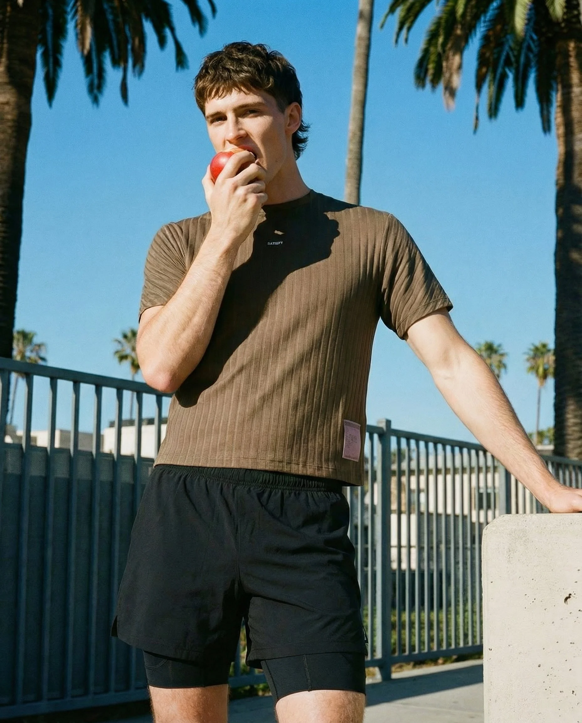 A young man in athletic clothing biting into a red apple outdoors near a black metal fence, with palm trees in the background on a sunny day.