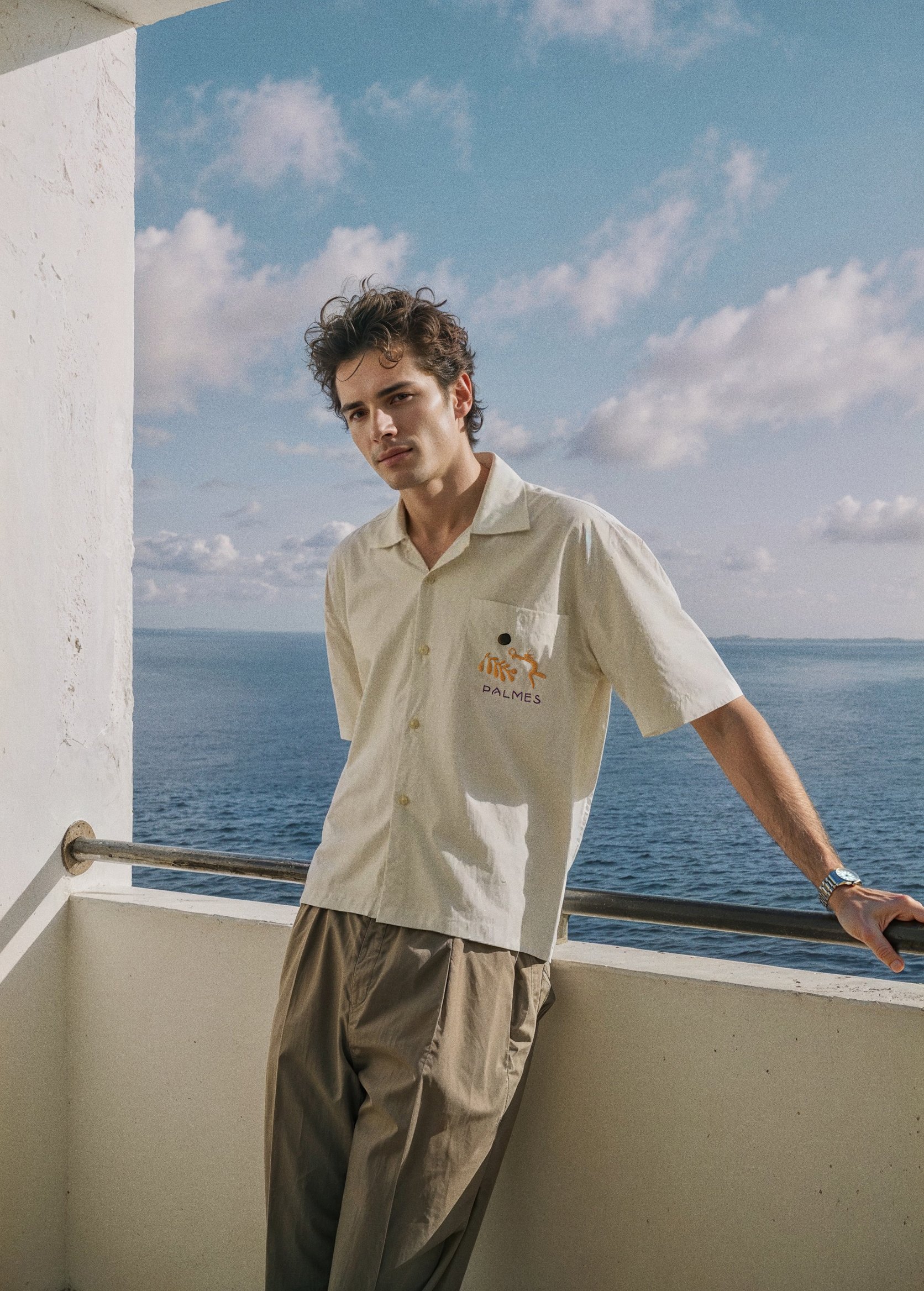 A young man leaning on a railing on a balcony overlooking the ocean. He is wearing a beige short-sleeved shirt with a palm tree logo and the word "PALMES" embroidered on it, and khaki pants. The sky is partly cloudy with a view of the water in the ba