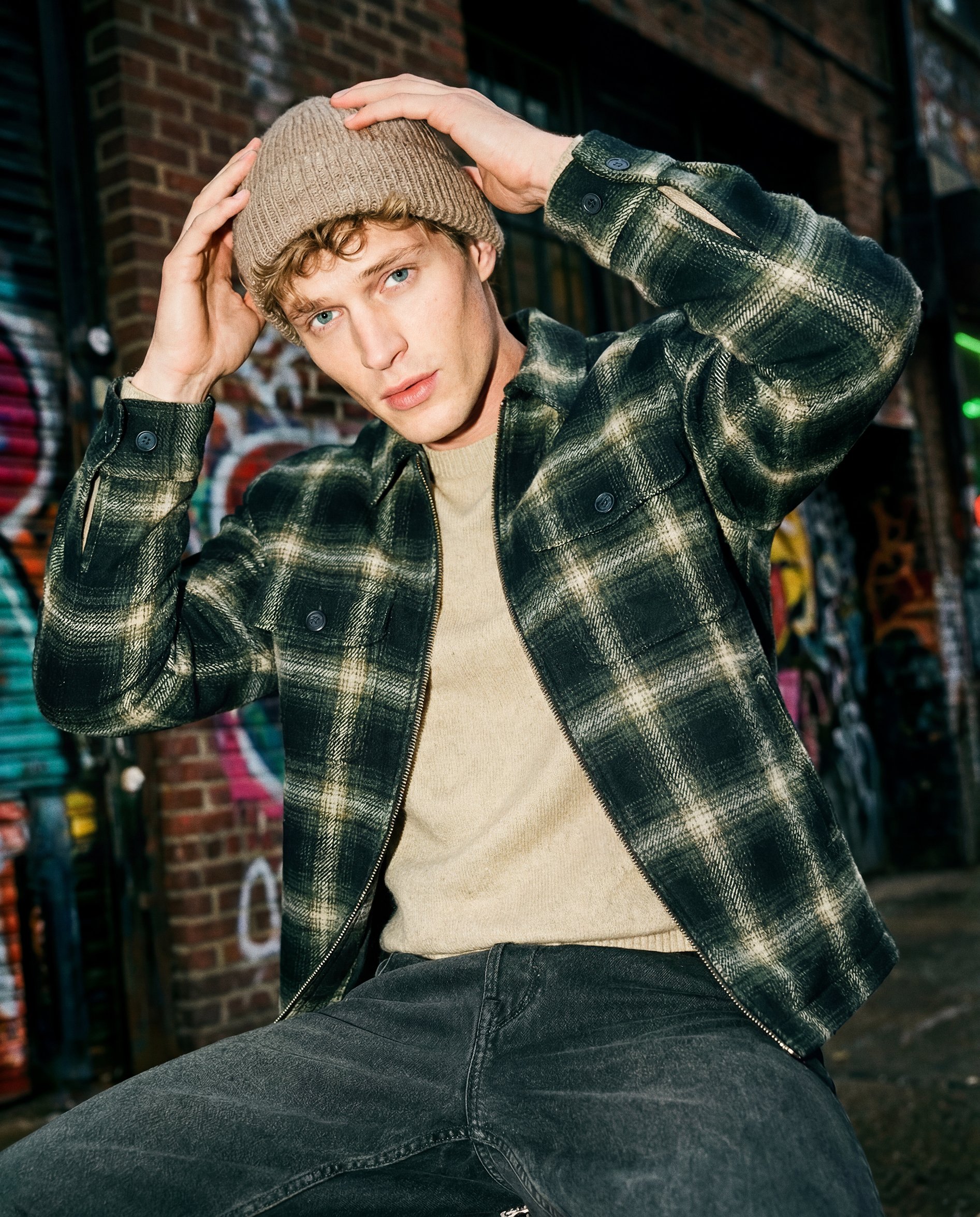A young man with blond curly hair and blue eyes is wearing a tan knit beanie, a beige sweater, and a green plaid jacket while sitting outdoors at night, with a background of a brick wall and shelves with colorful items.