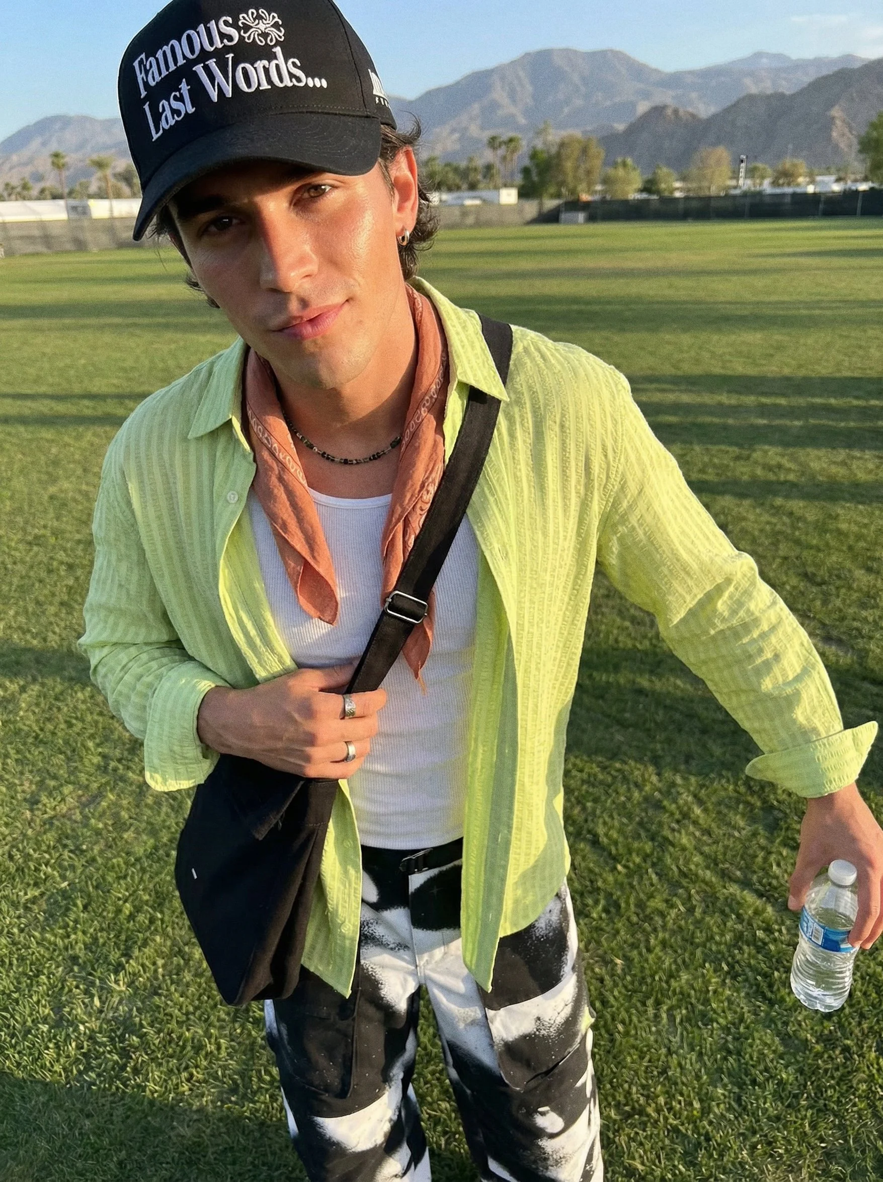 A young man standing on a grassy field with mountains in the background. He is wearing a black baseball cap with the text 'Famous Last Words...' and a yellow shirt over a white tank top. He has earrings, a necklace, and a ring, and is holding a water