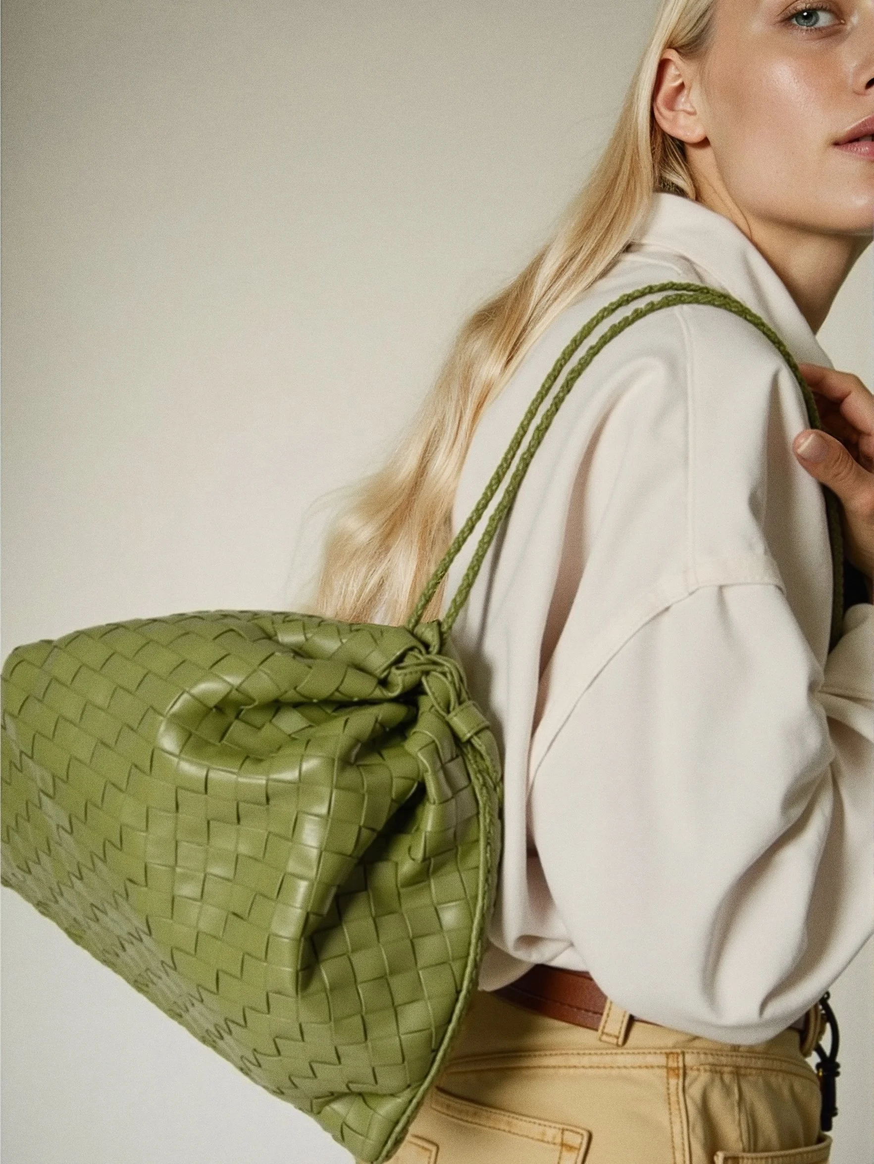 A woman with blonde hair wearing a beige jacket, tan pants, and a brown belt, carrying a green woven bag on her shoulder. The photo captures her from the side against a plain beige background. Brands: Bottega Veneta, Acne Studios Ai editorial image.
