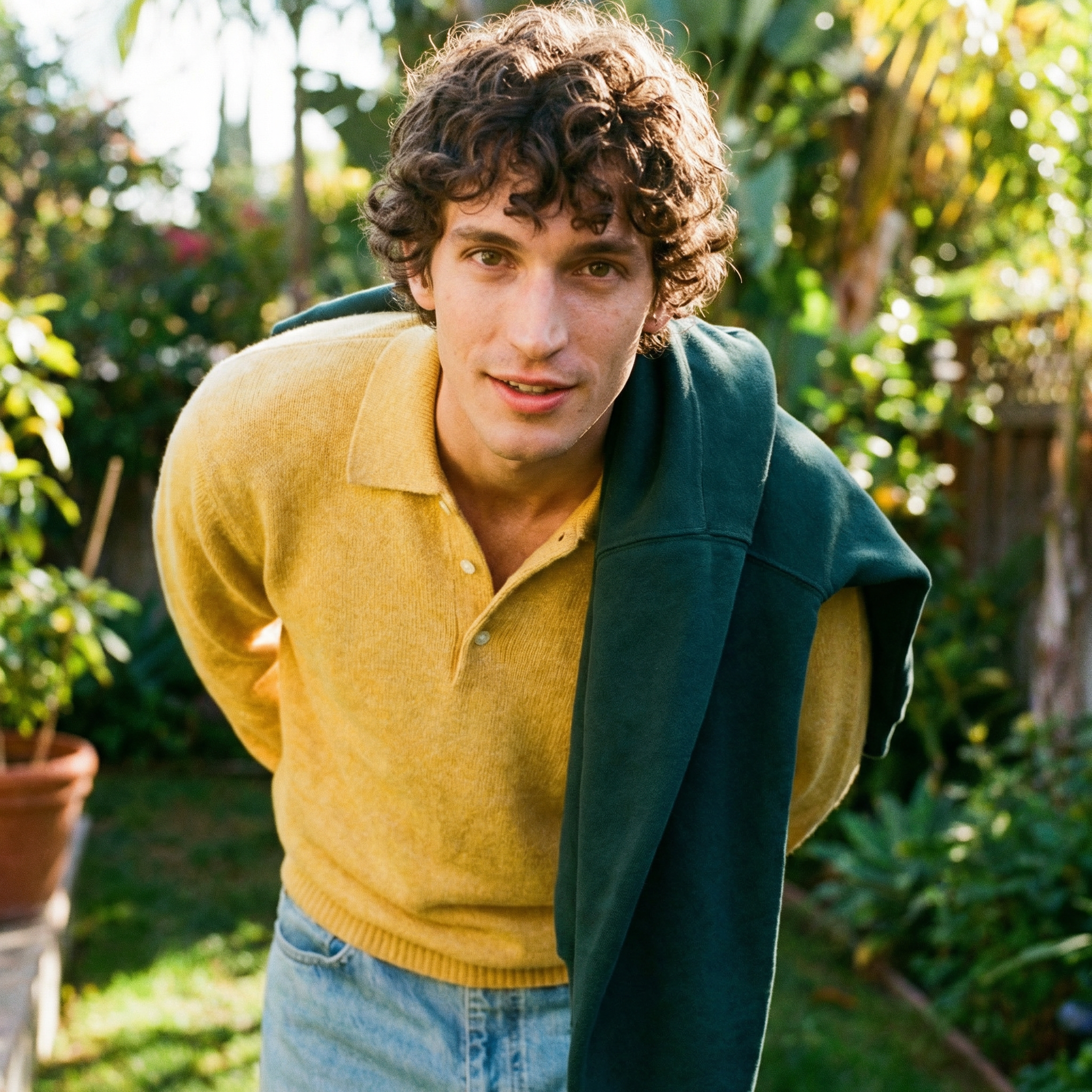 A young person with curly hair, wearing a yellow sweater and a green jacket, outdoors in a garden with lush greenery and sunlight.