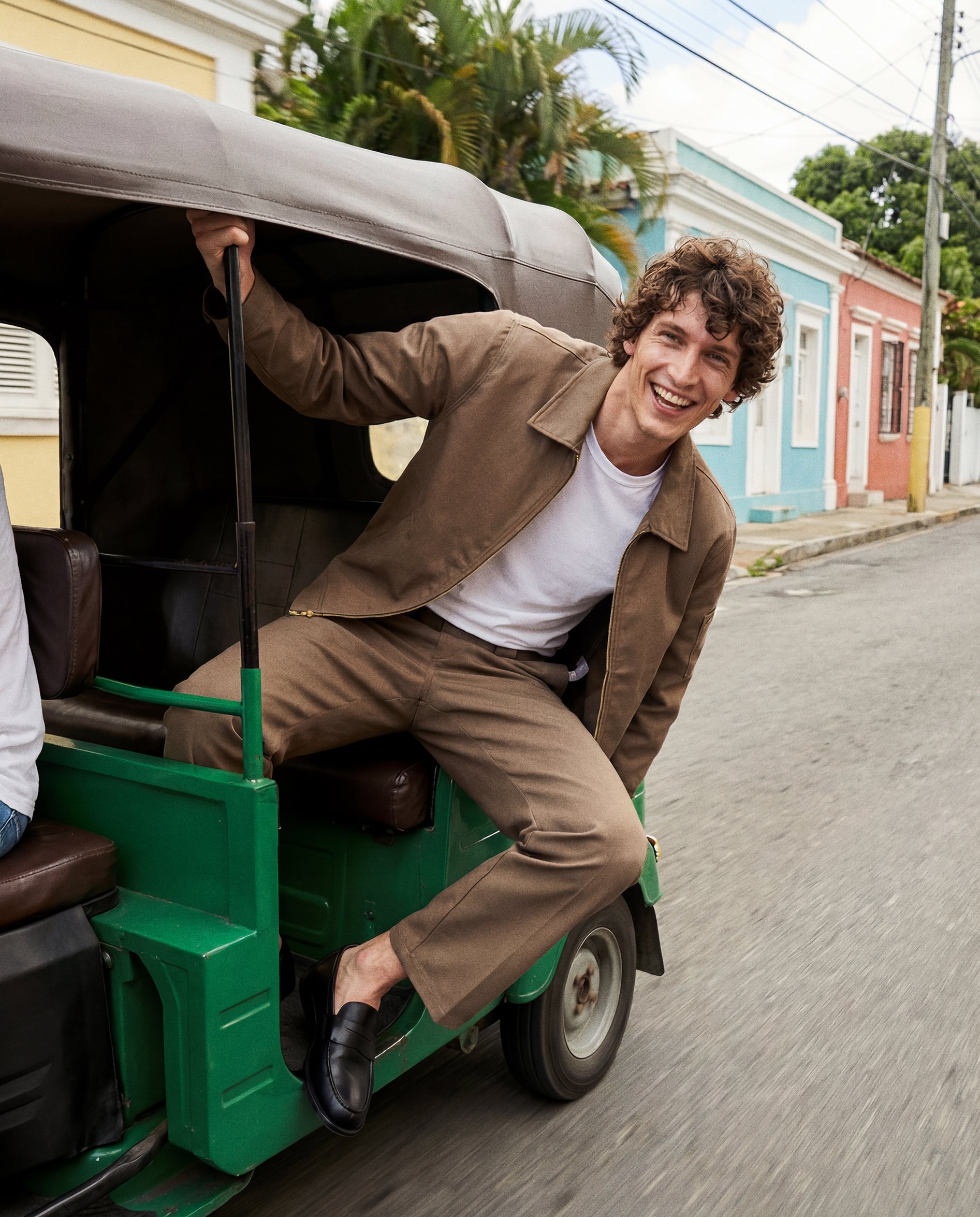 Young man with curly brown hair smiling and leaning out of a green golf cart on a street with colorful houses, palm trees, and power lines in the background.