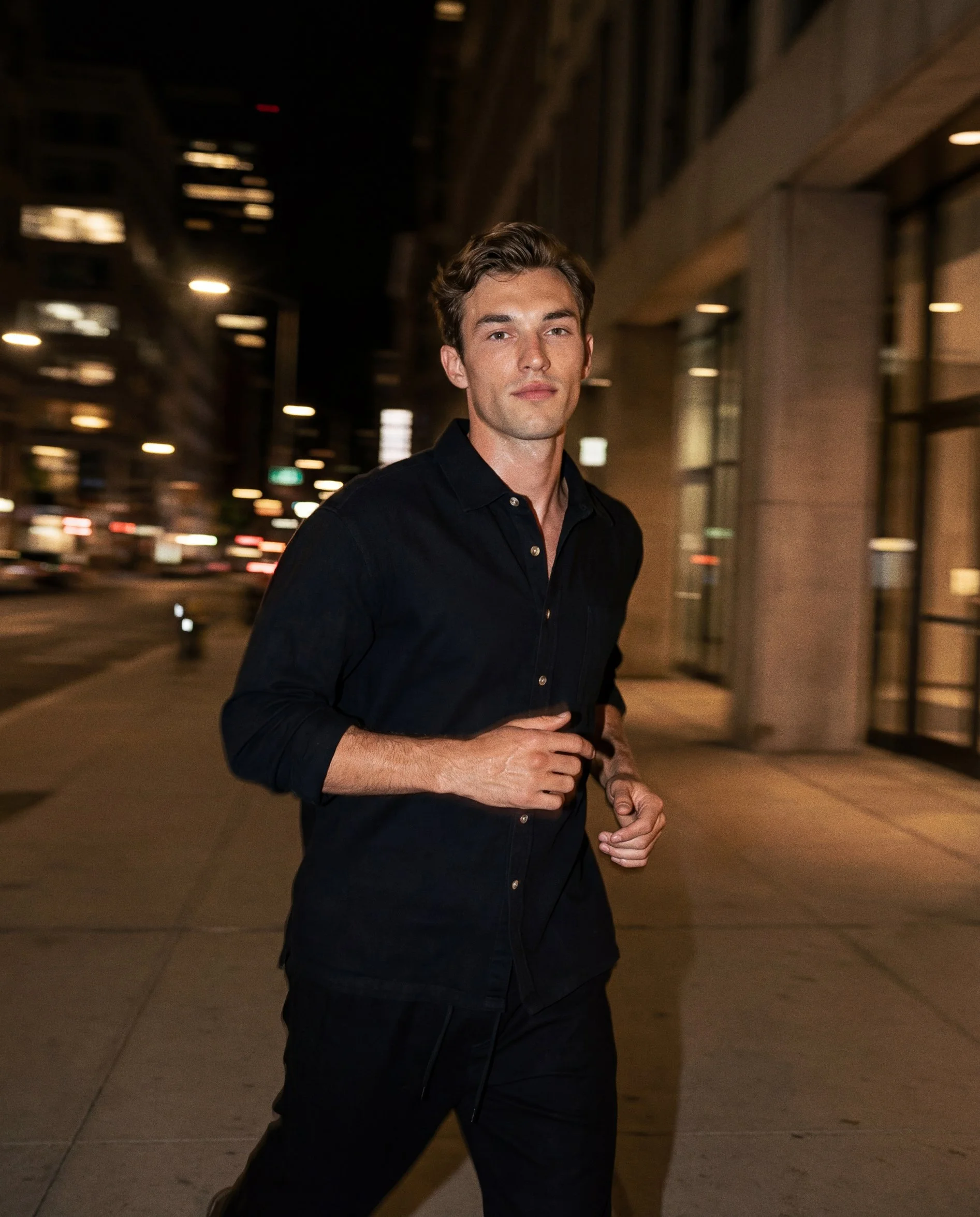 A young man with light skin, brown hair, and blue eyes walking on a city sidewalk at night, wearing a black shirt and black pants.